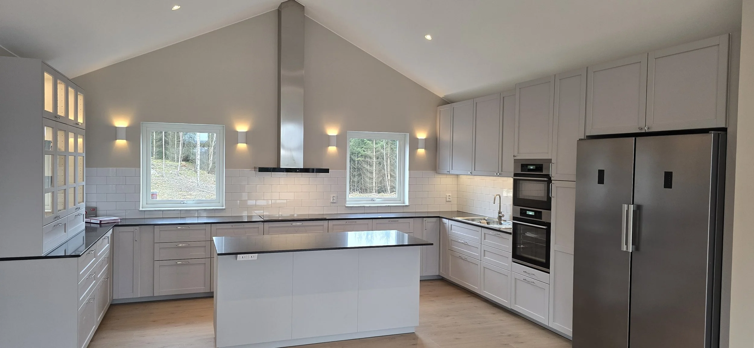 Modernt vitt kök med kökssnickerier, svart bänkskiva och rostfria vitvaror, inklusive stor kylskåp och inbyggda ugnar. Två fönster och vägglampor ger ljus till rummet.