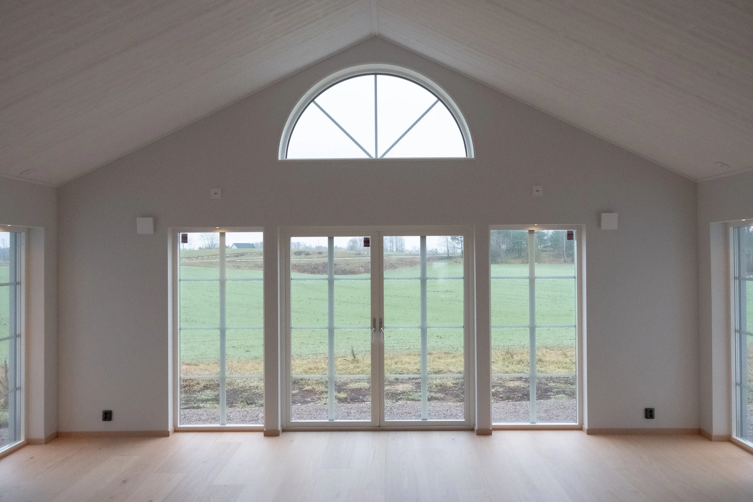 Ett tomt rum med stora fönster och utsikt över ett grönt landskap, med en halvskuggad trappa och ett vitt tak.