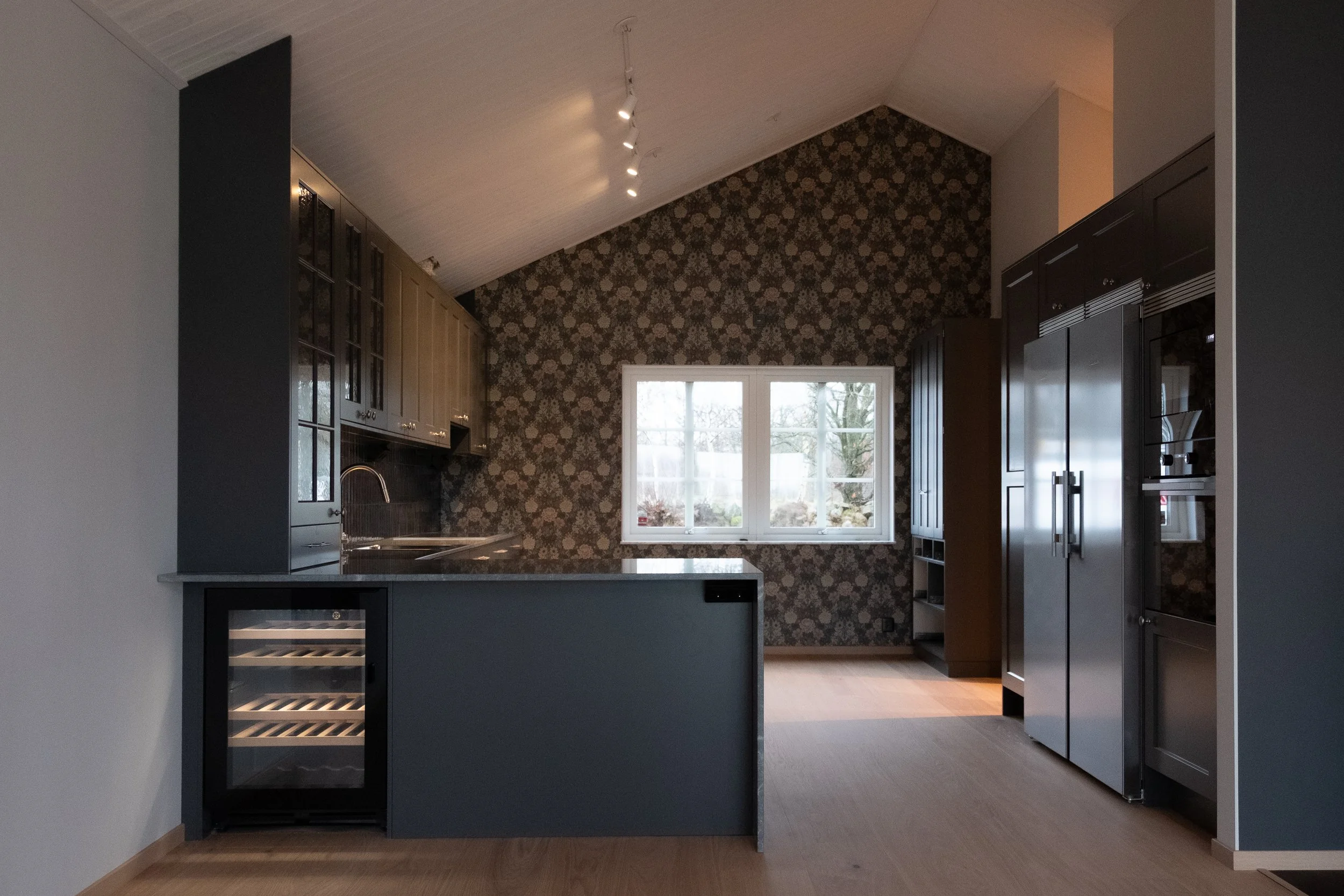 Mörkgrått kök med varmluftsugn, kylskåp, diskho och fönster mot trädgård.