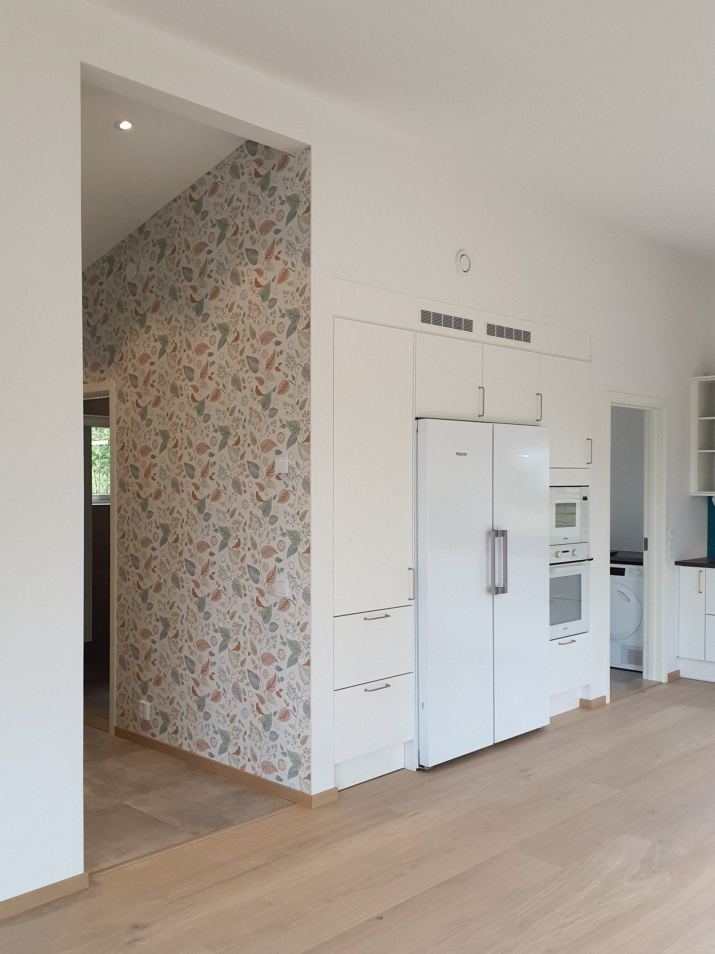 Vitt kök med inbyggda vitvaror och en vägg med färgglad blommig tapet.