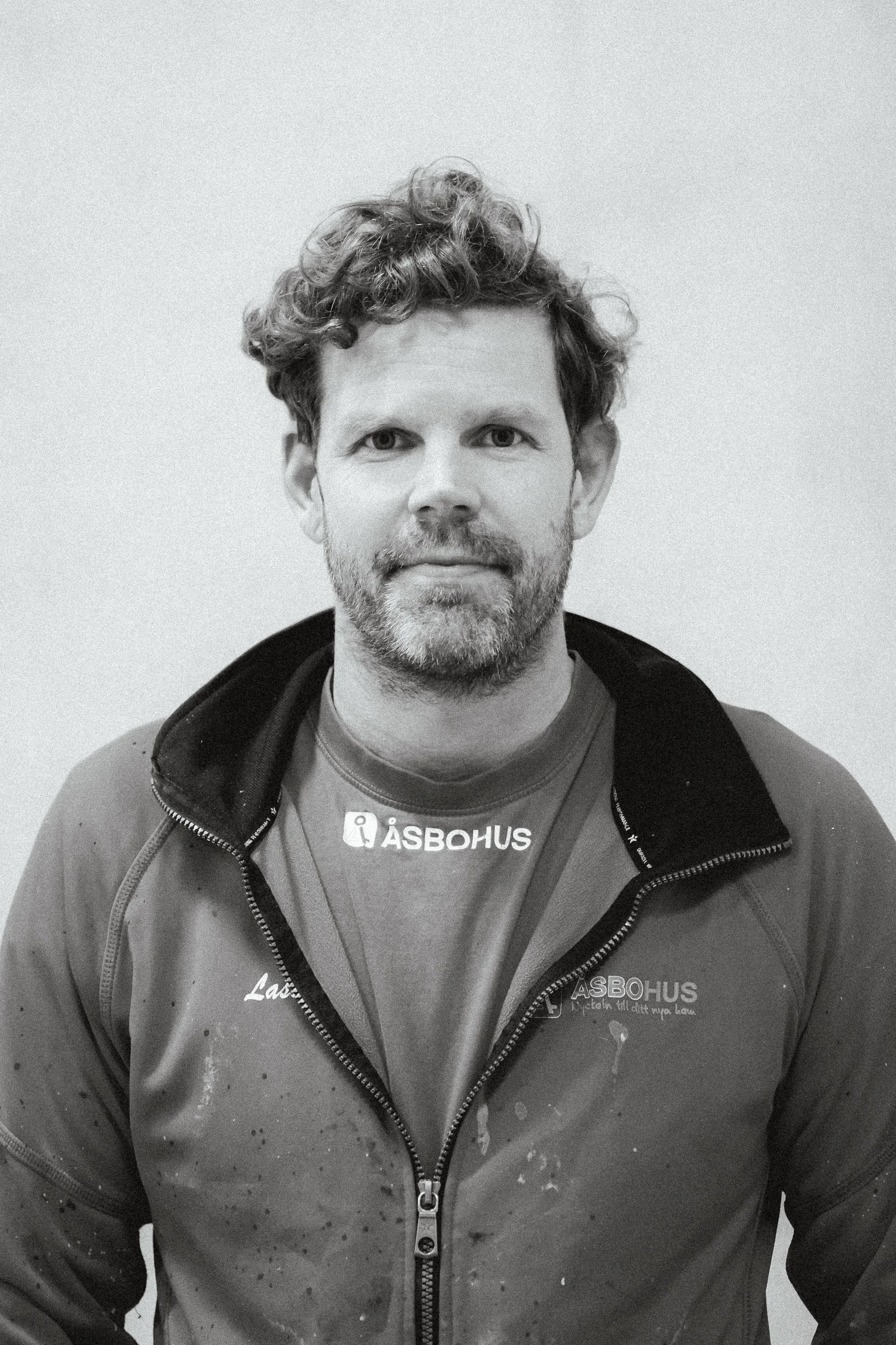 Person i sportjacka med logotypen åsbohus, står framför en ljus vägg, svartvitt foto, kort hår och skägg.