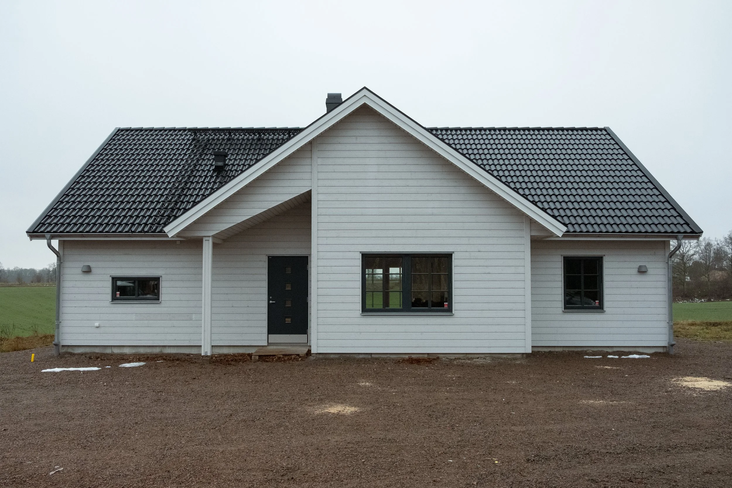 Vit trästuga med svart tak och tre fönster, omgivet av öppen mark och gräs i bakgrunden.