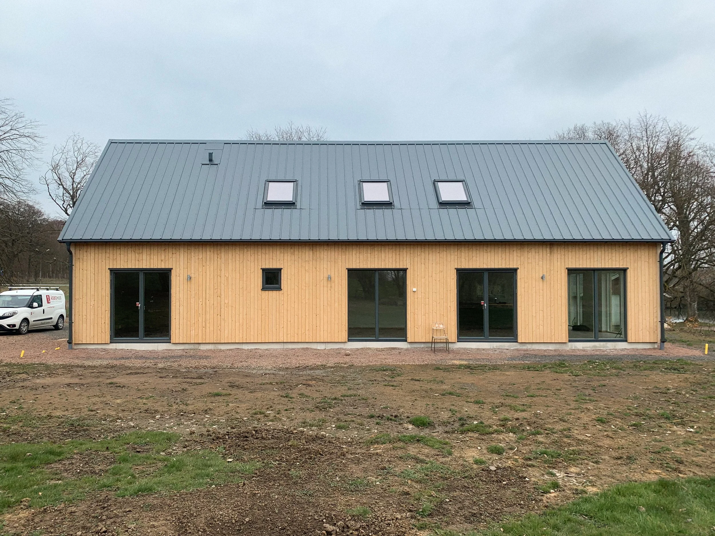 Nyrenoverad modern hus med gul träfasad och svart tak, fyra stora glasdörrar mot trädgården, tre takfönster och två mindre väggfönster, utomhusområde med jord och gräs.