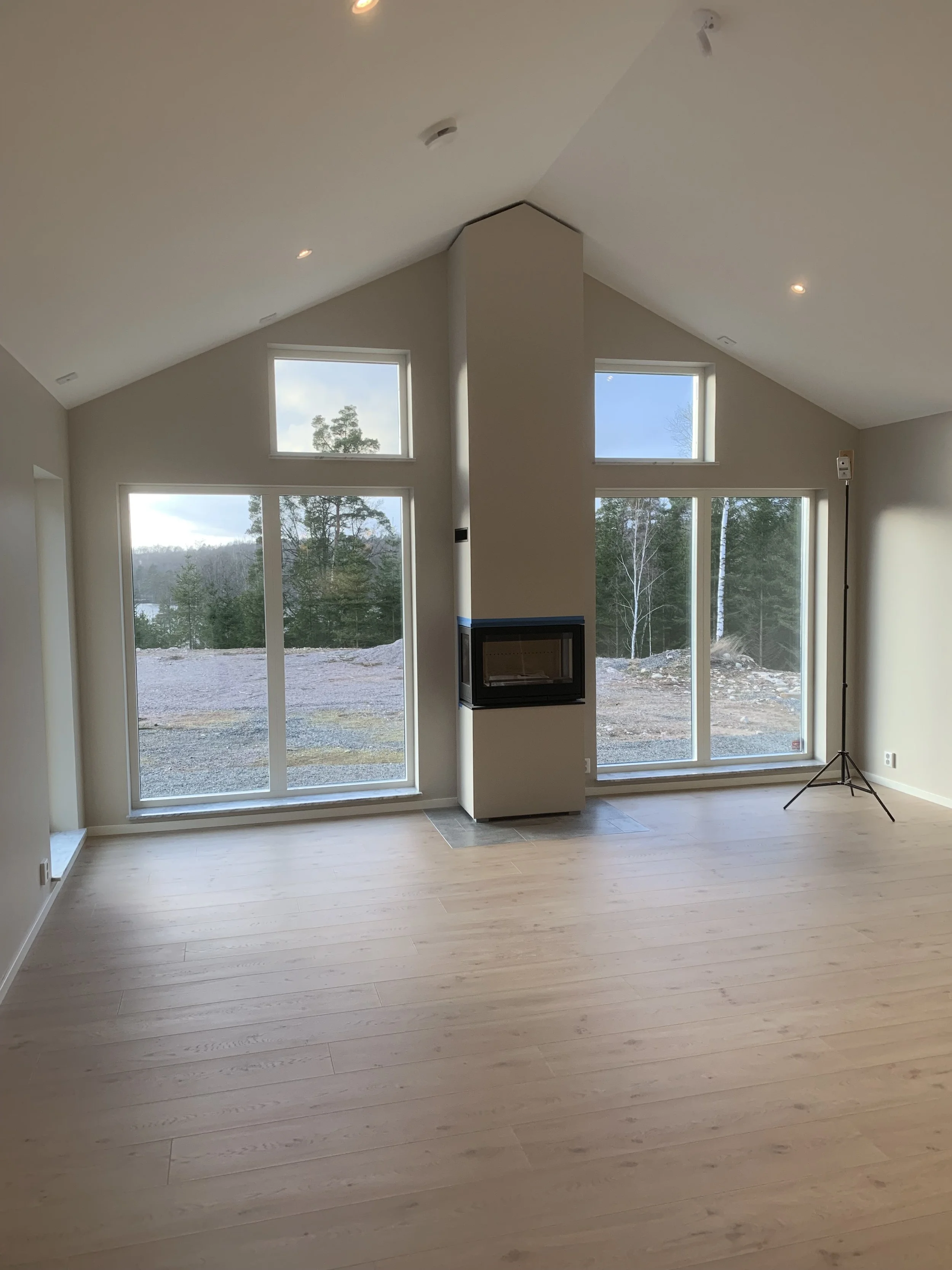 Ett tomt vardagsrum med stora fönster, en väggfast öppen spis, ljust trägolv och omgivande landskap utanför.