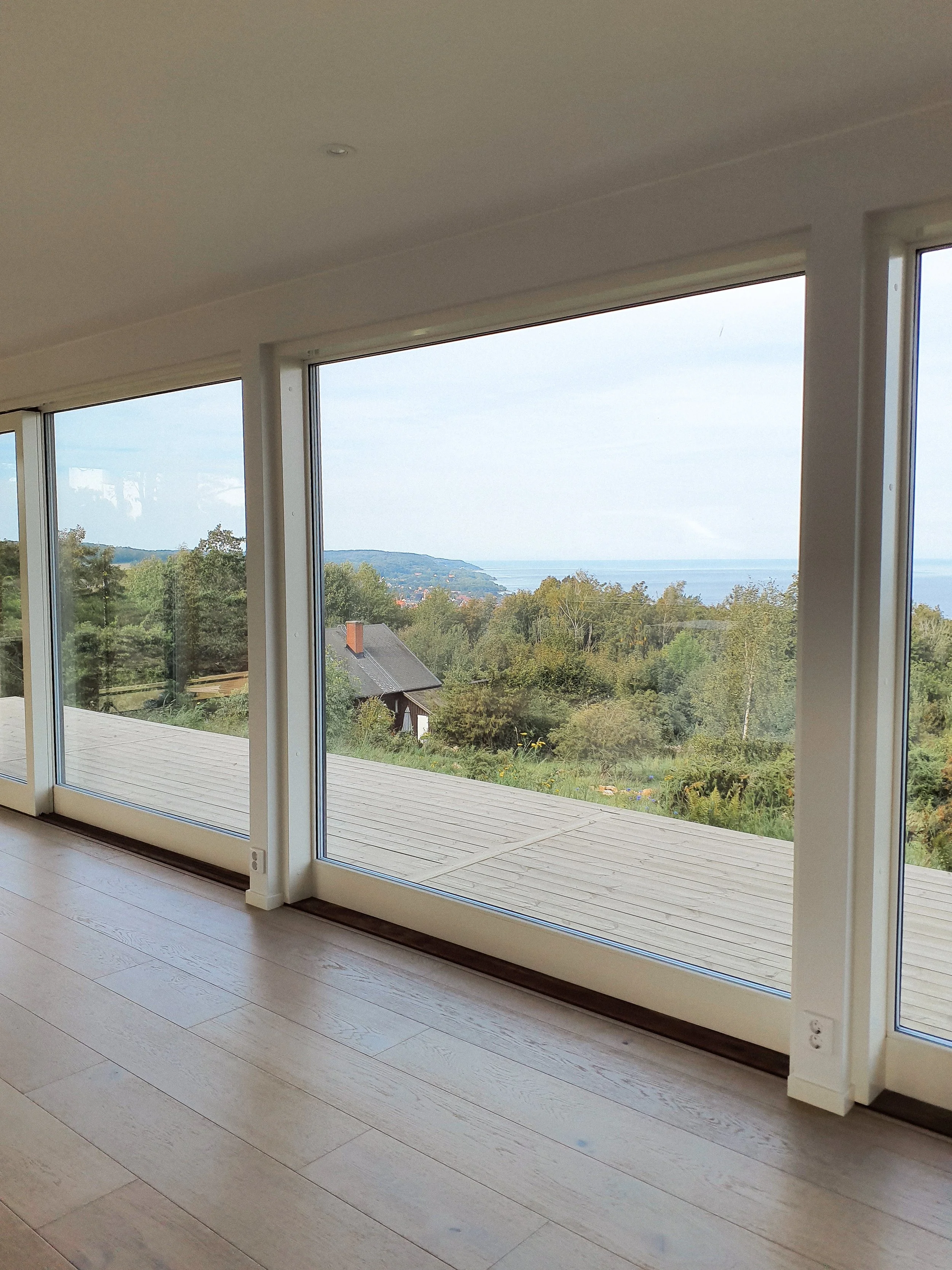 Interiör med stora fönster som visar en grön landskap med träd och en kropp av vatten i bakgrunden.