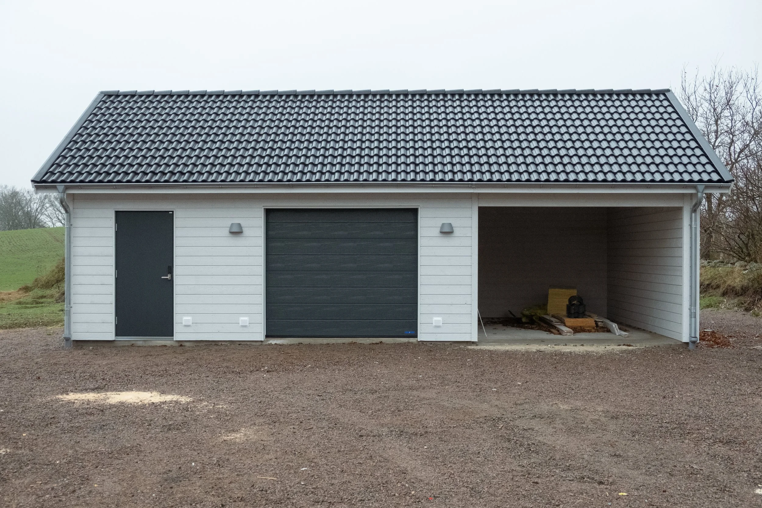 En vit liten garagebyggnad med två dörrar och ett öppet utrymme till höger på en grusig mark, omgiven av träd och gröna landskap.