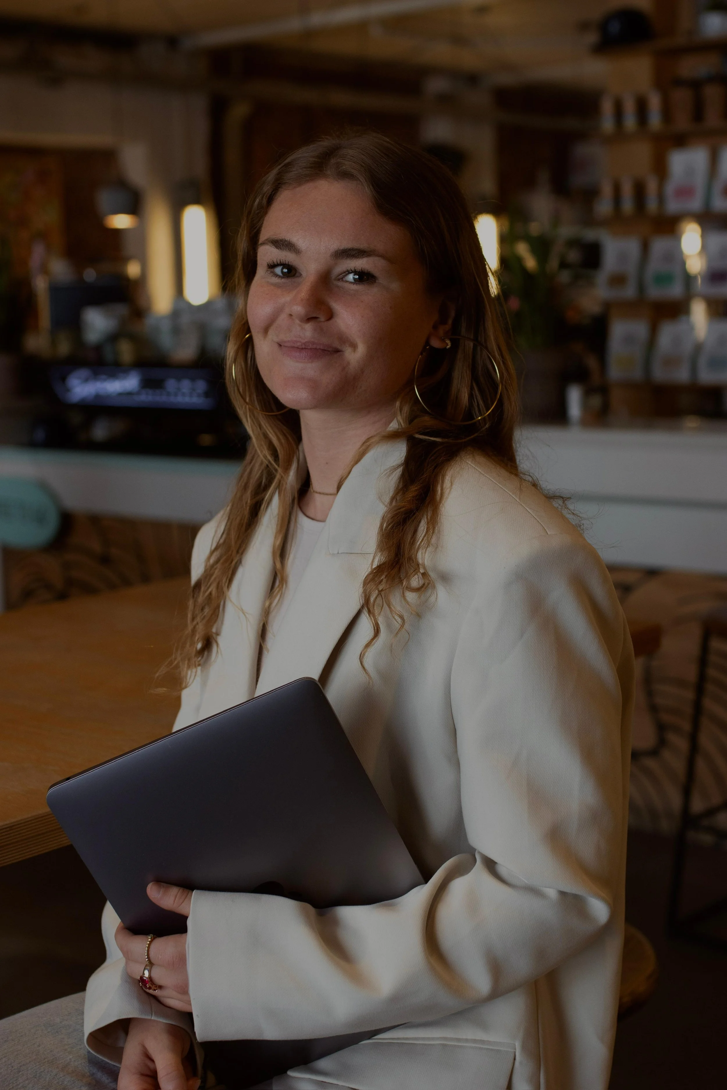 Een jonge vrouw met bruin haar draagt een beige jas en houdt een donkerkleurige map vast. Ze staat binnen in een gezellige ruimte met houten interieur en schapenkast achter haar. Branding shoot
