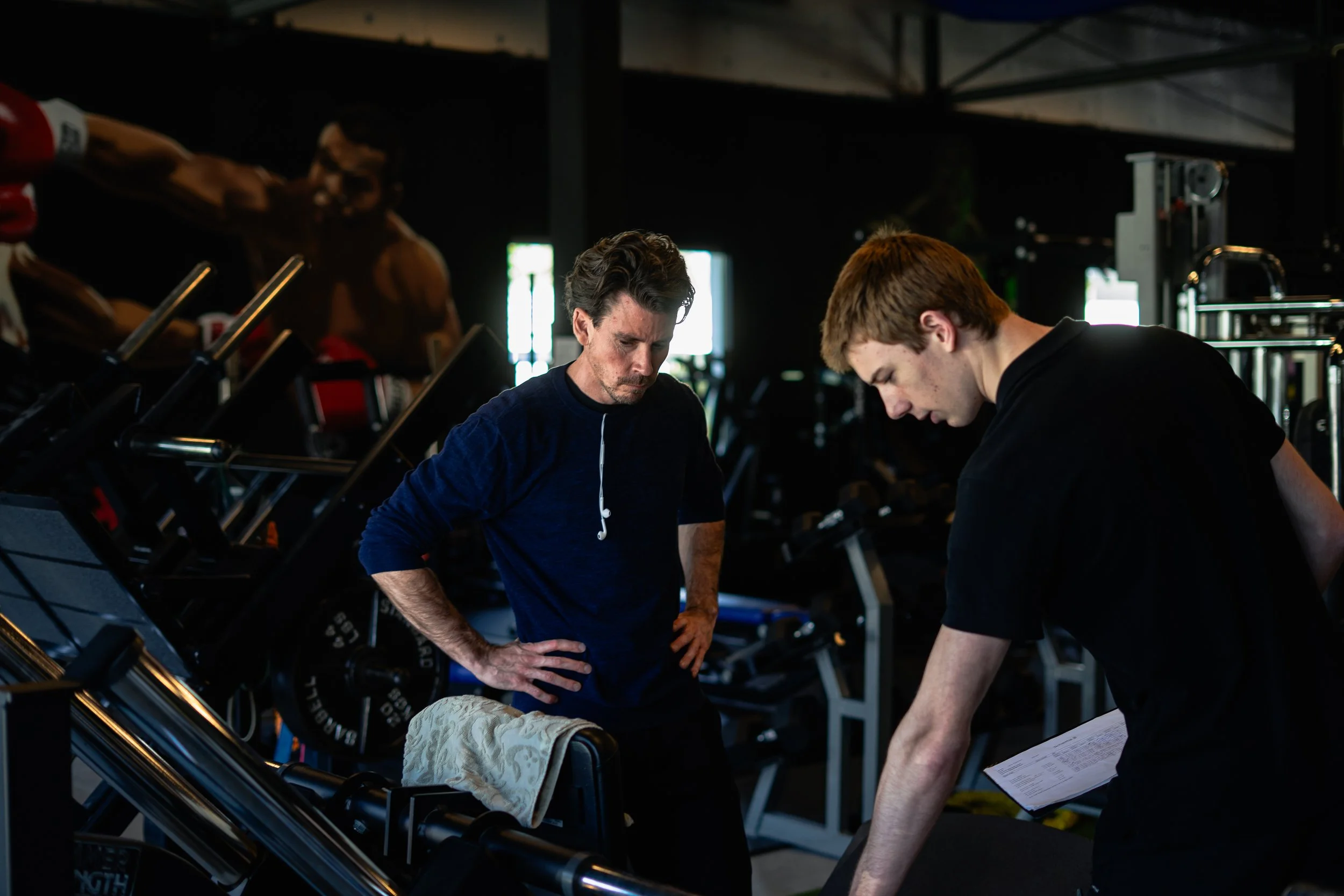 Twee mannen in een fitnessruimte, de ene kijkt serieus en heeft zijn handen op zijn heupen, de ander houdt een stuk papieren in zijn hand en luistert. Er is fitnessapparatuur op de achtergrond. Sportschool Emmen. Krachttraining.
