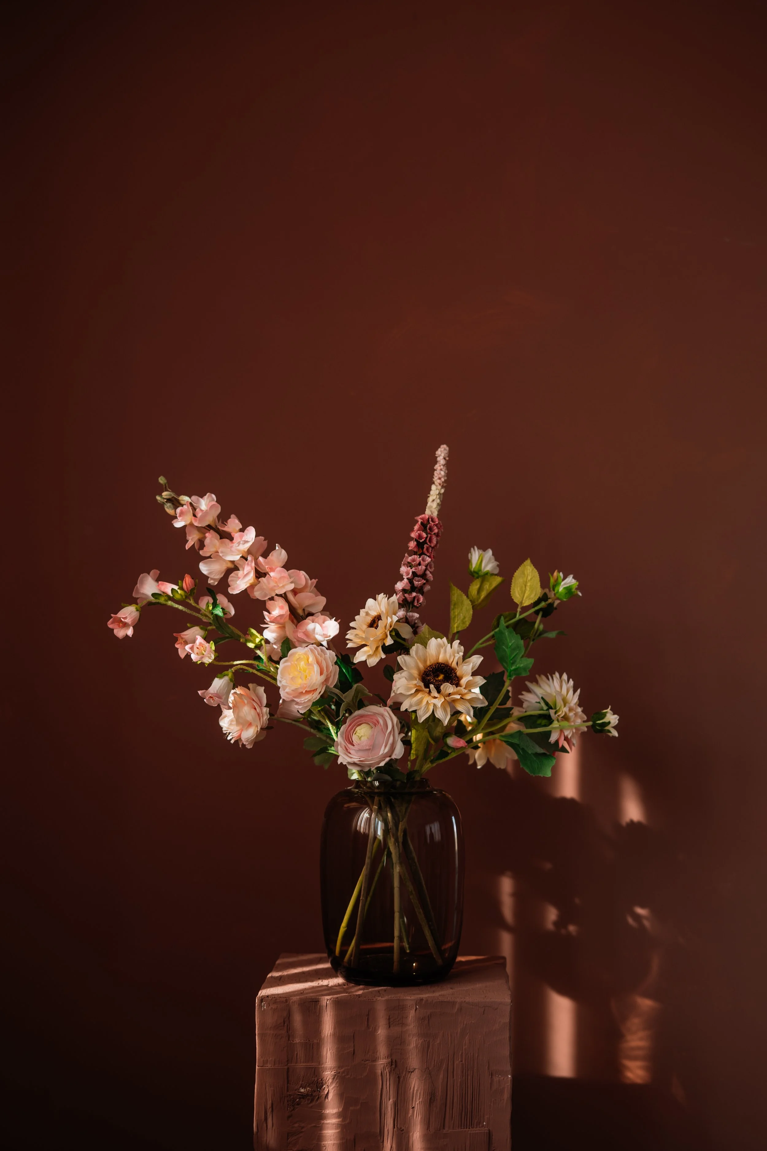Een boeket van diverse bloemen in een donker glas vazen op een houten sokkel, met een donkere achtergrond. Floweris