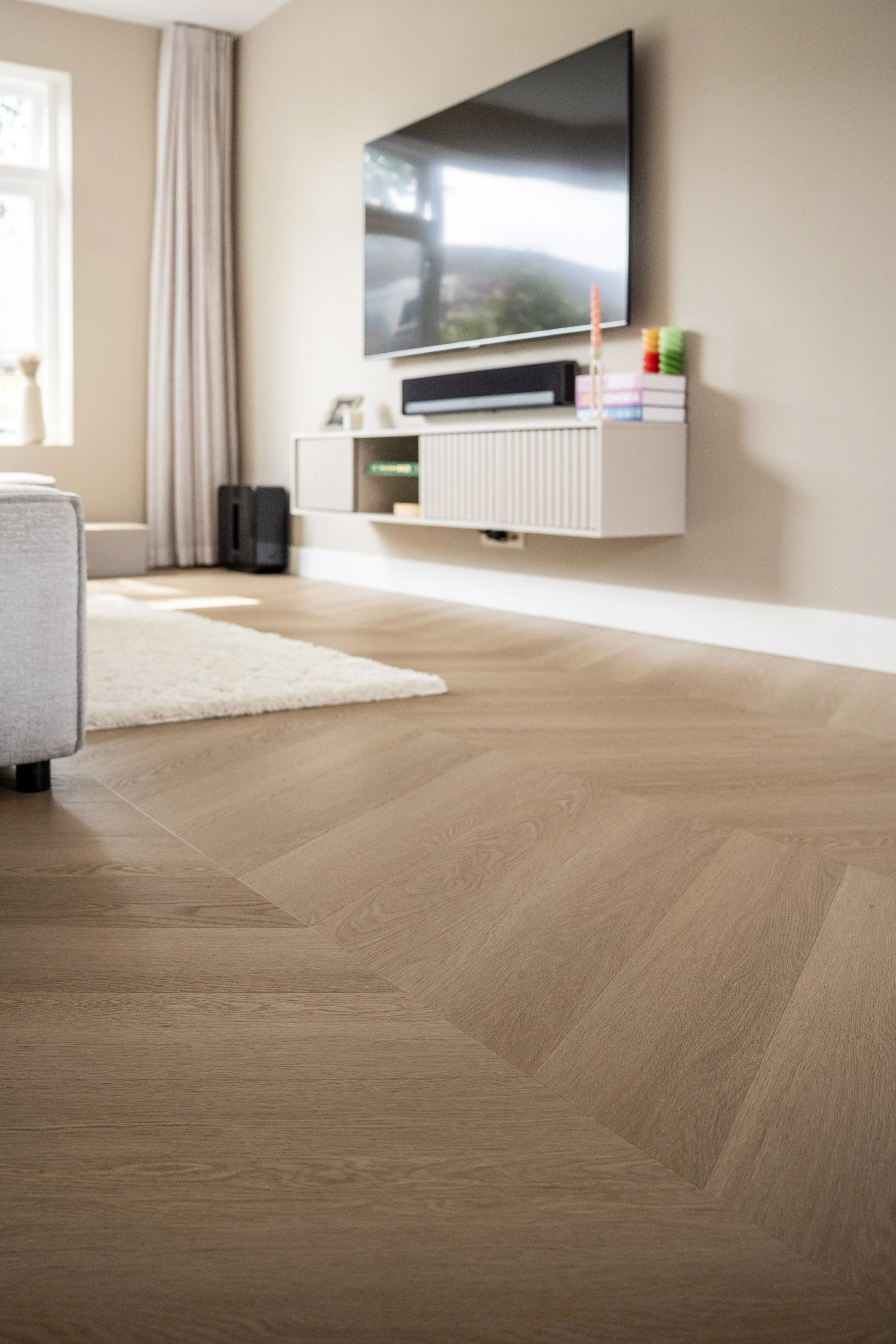 Moderne woonkamer met houten vloer, wandmeubel, grote televisie en raam met beige gordijnen. Laminaatpaleis Ede