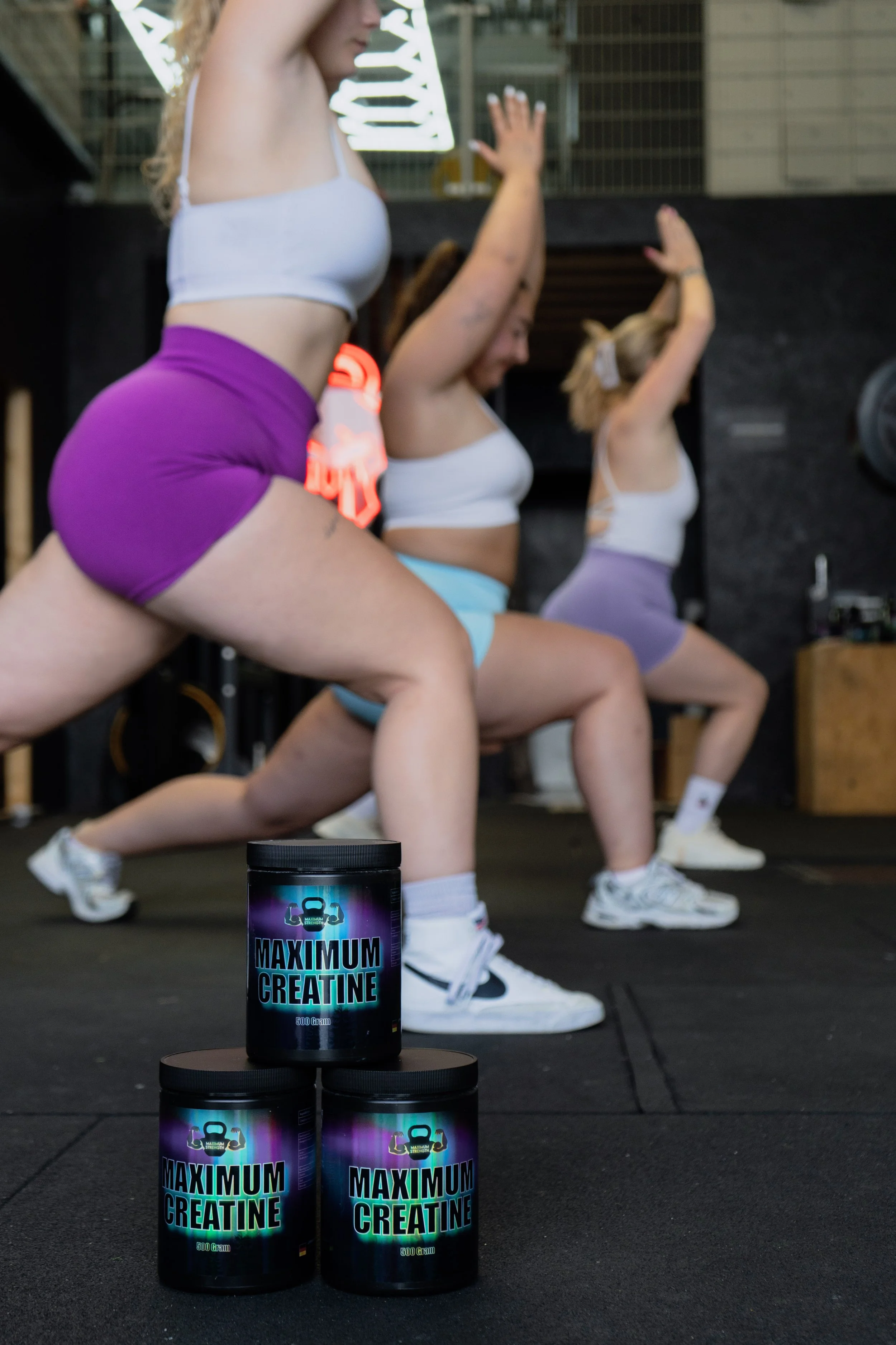 Drie vrouwen die in een sportschool squats doen, met drie potten Maximum Creatine in de voorgrond.