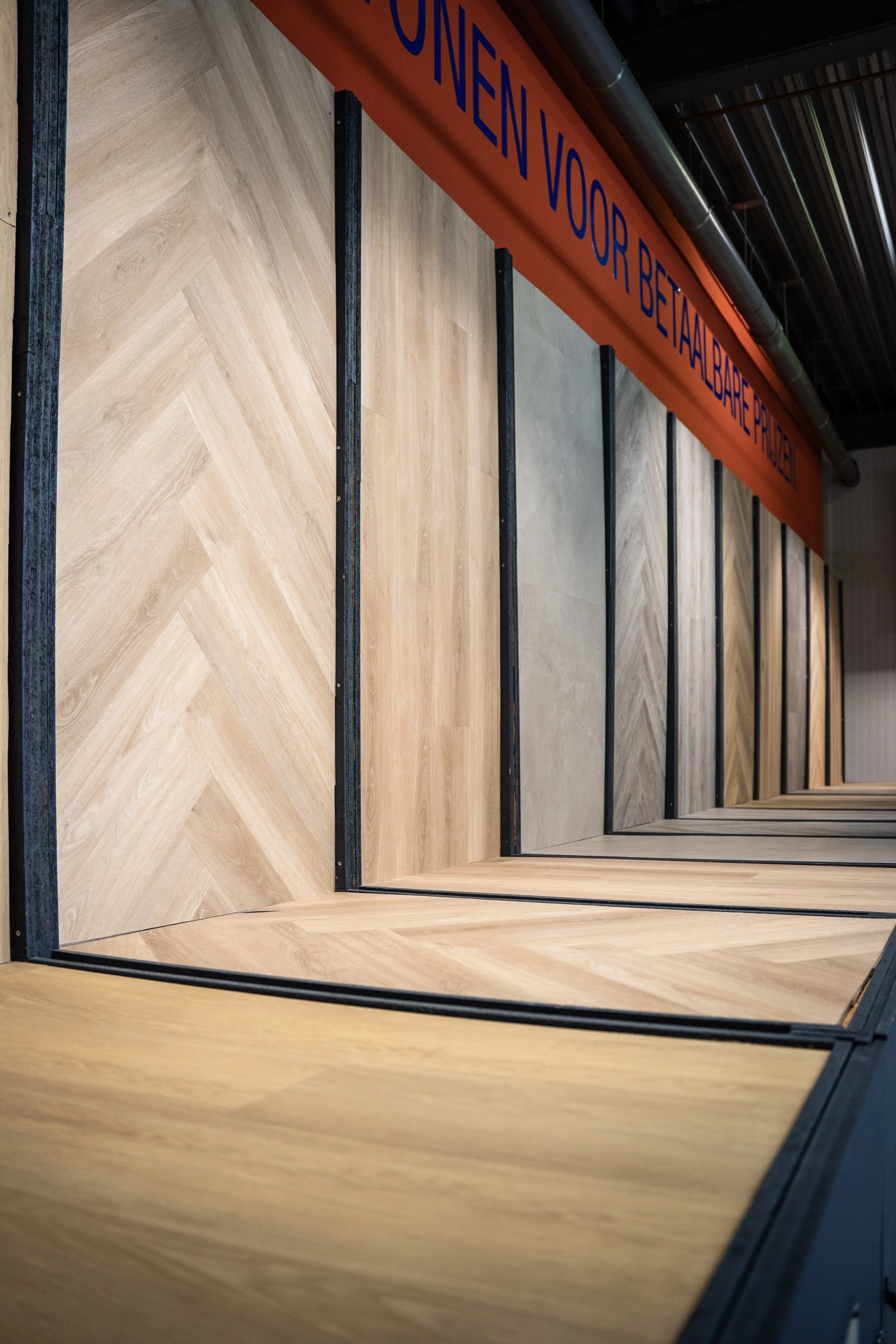 Foto van verschillende houten vloerpanelen gezet in een winkel of showroom, met een rood bord met blauwe tekst erboven. Laminaatpaleis Ede