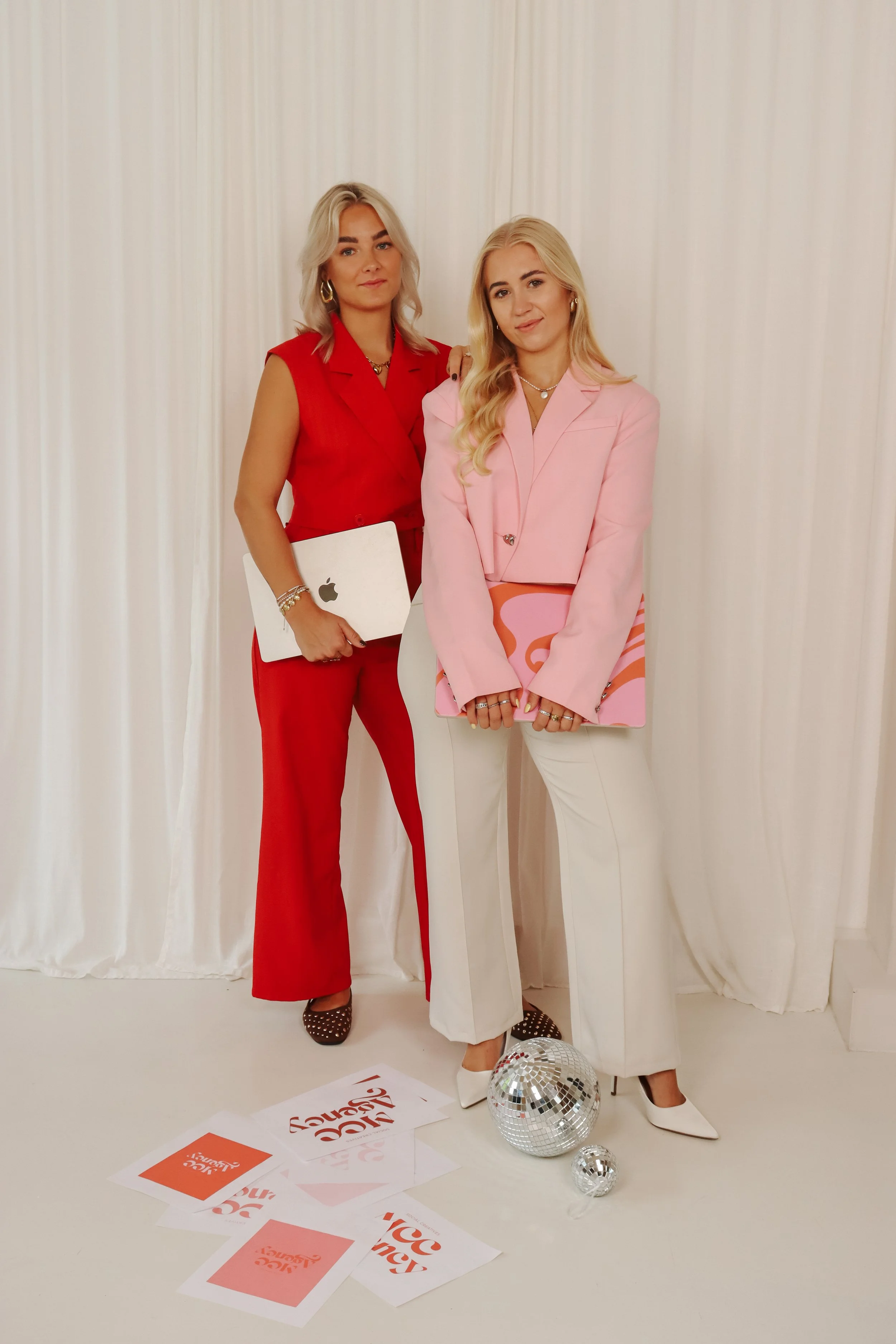 Twee vrouwelijke fashionista's poseren voor een witte fluwelen achtergrond. Ze dragen chique outfits, één in rood en de andere in pastelroze en wit, elk met accessoires en laptops. Op de vloer liggen beschikbare documenten en twee discobollen.