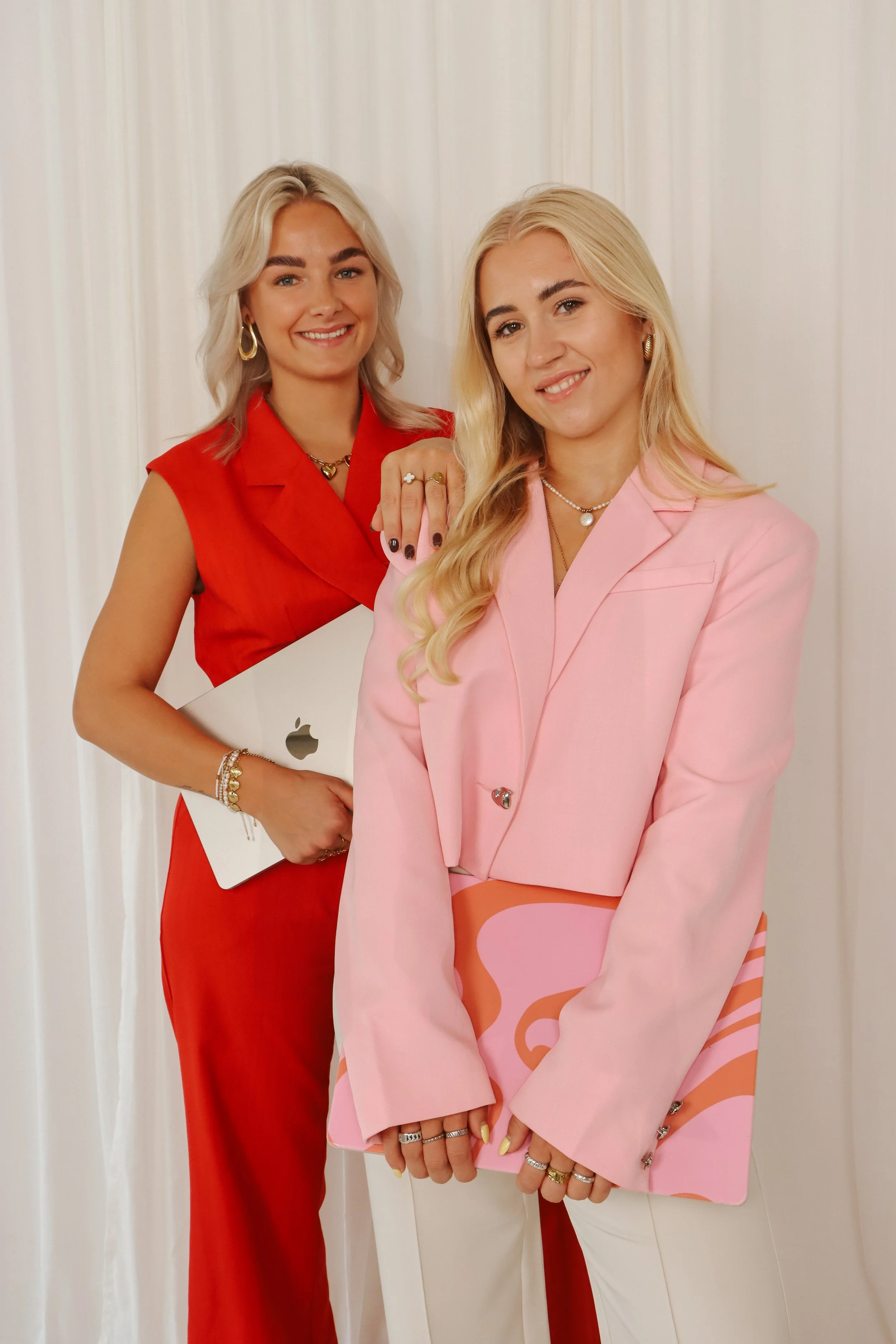 Twee jonge vrouwen, één met blond haar in een roze blazer en de ander met lichtblond haar in een rode jurk, poseren voor een witte achtergrond, beiden glimlachend.