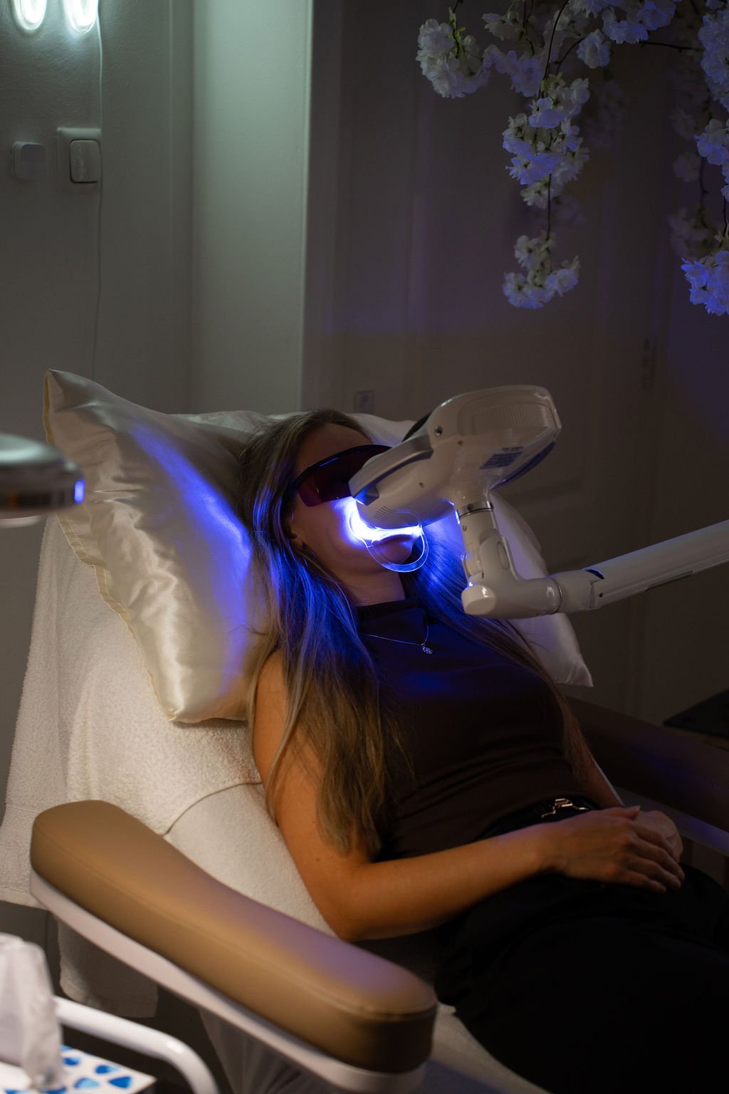 Een vrouw ligt in een onderzoekbed met een laserbehandeling op haar gezicht, terwijl ze een beschermende bril draagt. Er hangen bloemen aan de muur op de achtergrond.