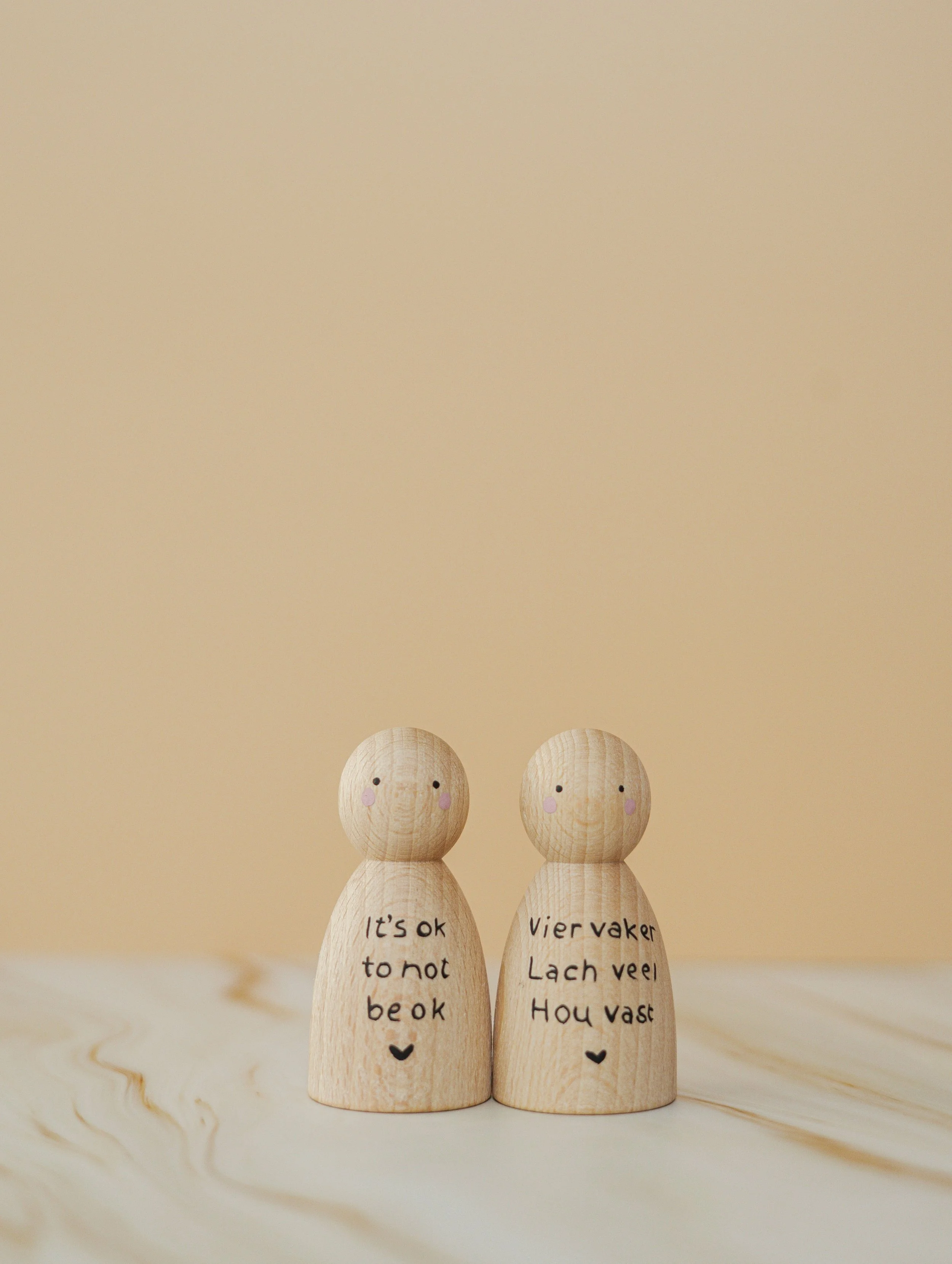 Twee houten figuurtjes met tekst en gezichtjes, op een houten oppervlak. De linker zegt 'It's ok to not be ok' en de rechter 'Vier vaker Lach veel Hoe vast'.