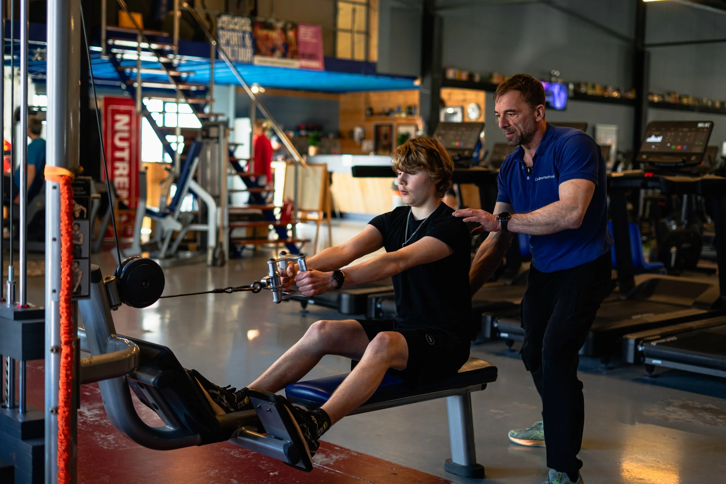 Een jonge jongen voert een krachttraining uit met een kabelmachine onder begeleiding van een coach in een sportschool.