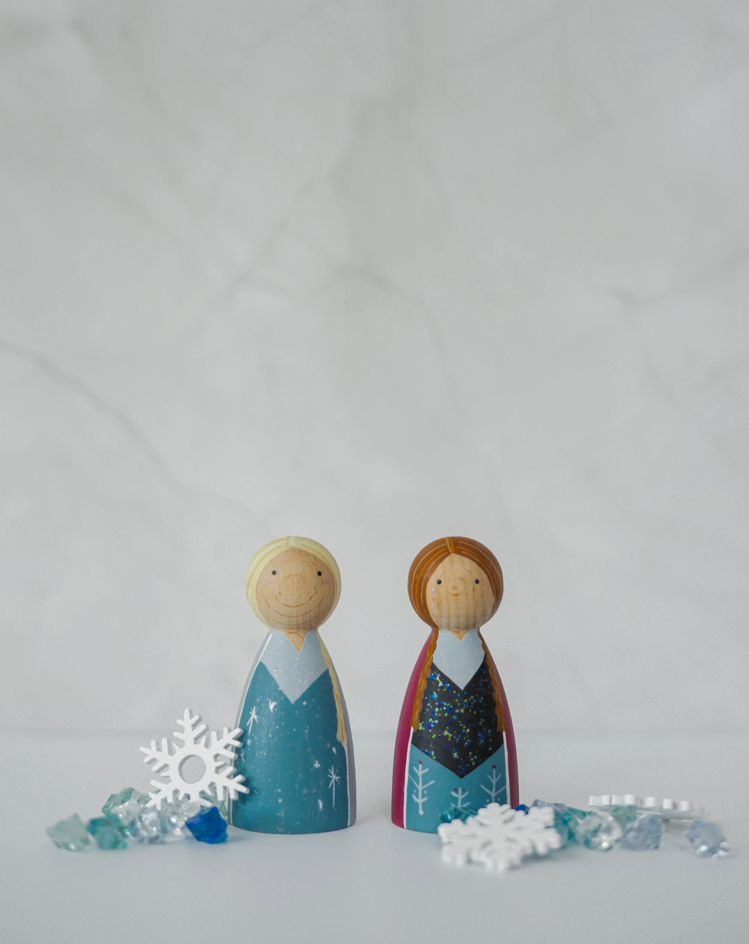 Twee houten kerstfiguren, een met een blauwe jurk en een met een zwarte en rode jurk, staan op een witte ondergrond met decoratieve sneeuwvlokken en kristalachtige decoraties eromheen.