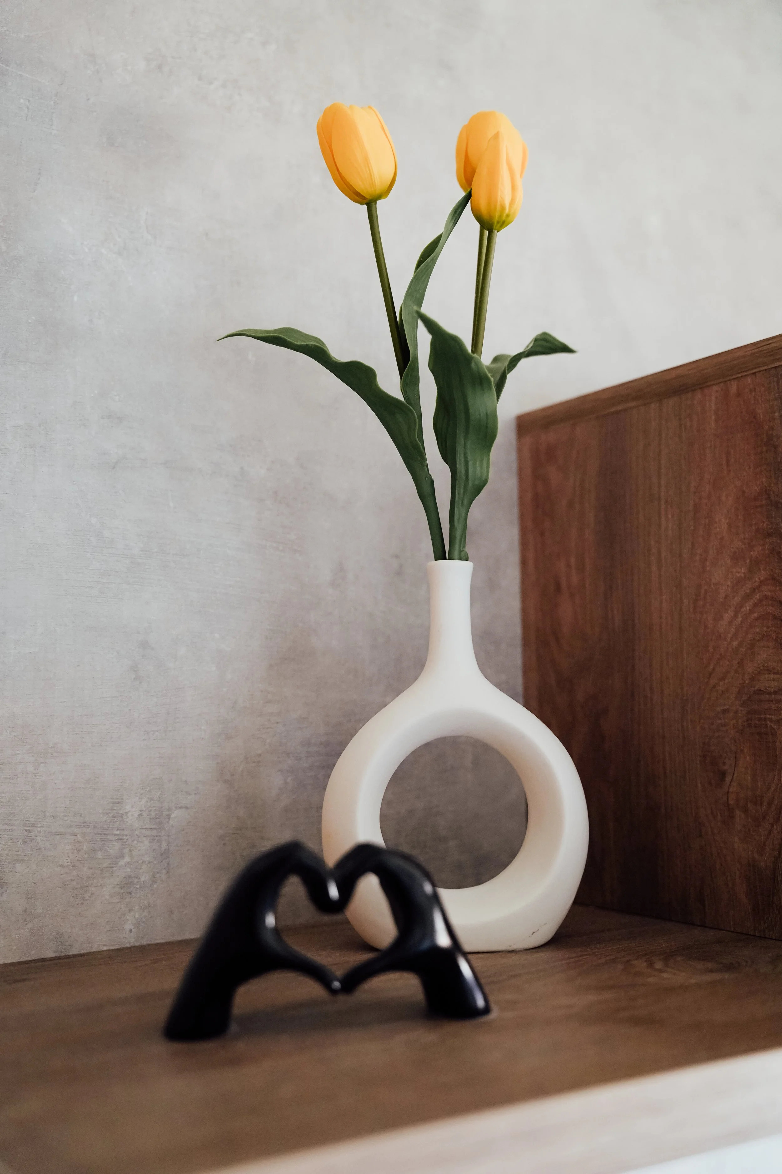 Een witte vaas met een cirkelvormig handvat bevat gele tulpen, geplaatst op een houten oppervlak. In de voorgrond ligt een zwart sculptuur in de vorm van twee handen die een hart vormen. Vakantiepark. Chalet.