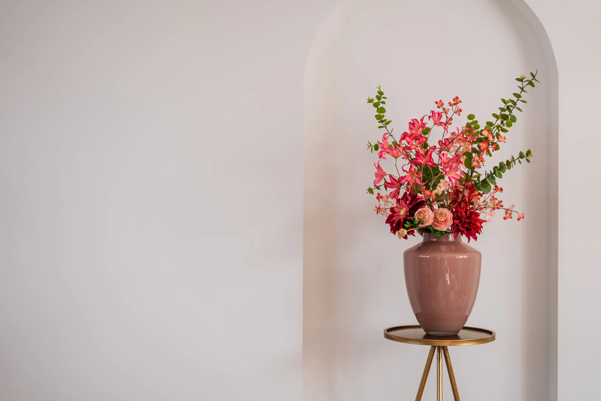 Een bloemstuk met roze en rode bloemen, gepresenteerd in een grote, roze keramieken vaas, geplaatst op een houten bijzettafel tegen een witte muur met een afgeronde nis. Floweris