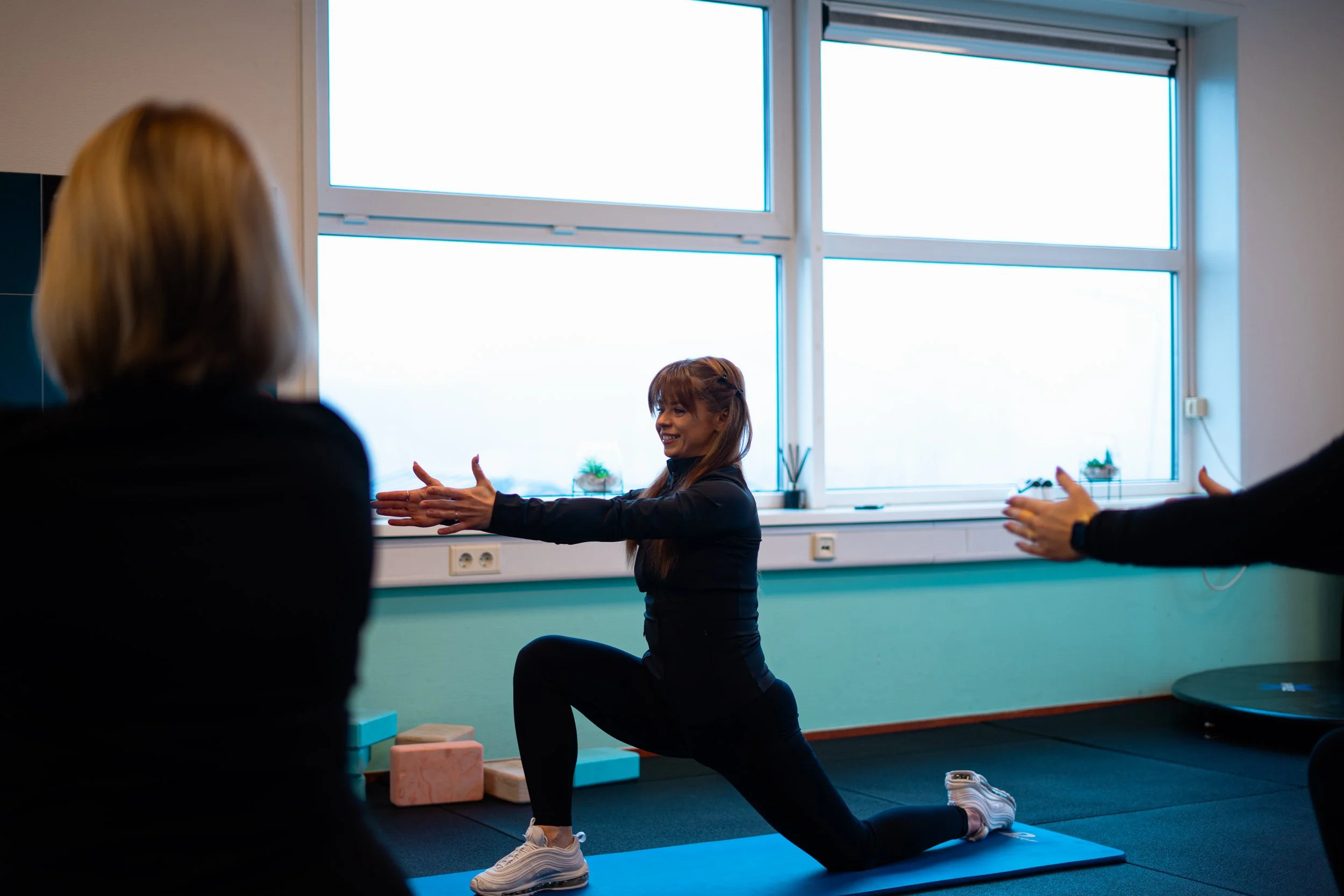 Vrouw die een yoga-oefening doet in een fitnessruimte, terwijl twee mensen op de achtergrond haar begeleiden.