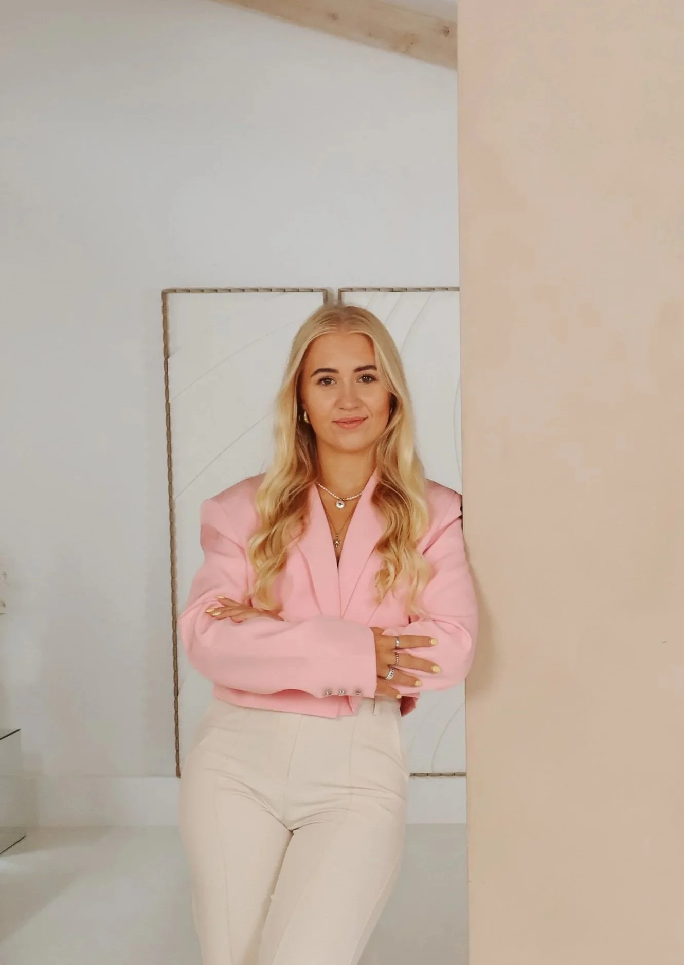 Vrouw met lange blonde haar, draagt een roze blazer en beige pantalons, staat met gekruiste armen voor een witte muur met decoratieve panelen. Branding shoot