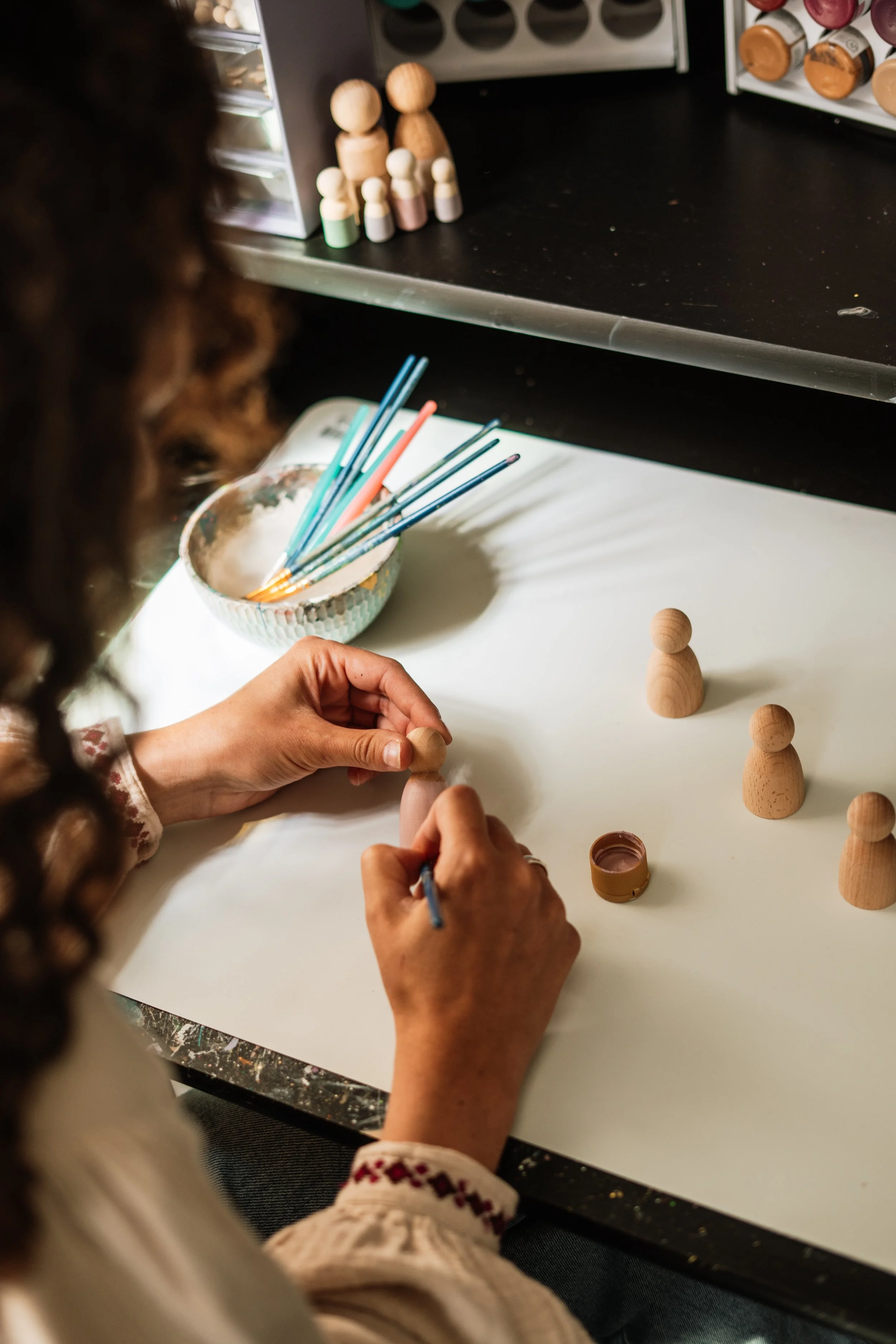 Een persoon schildert houten poppetjes met roze verf, naast een kom met verfkwasten en een plankenrek met verfpotten.