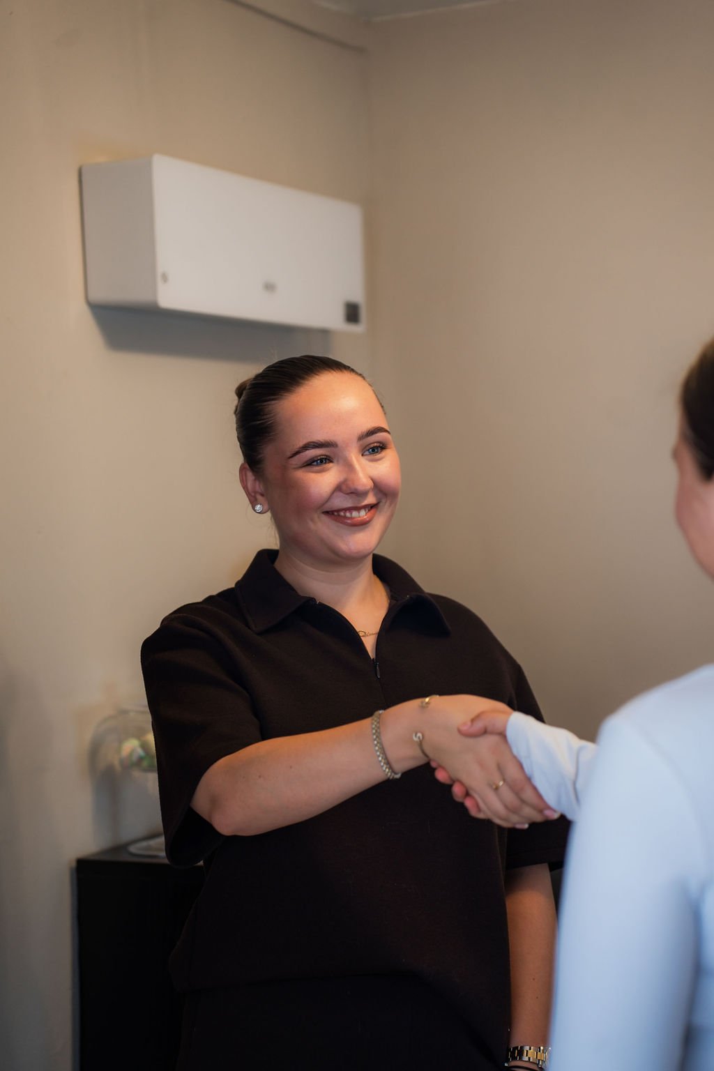 Twee mensen schudden handen, één vrouw met donker haar en een zwarte blouse, lachend, binnen in een kamer. Beautysalon.