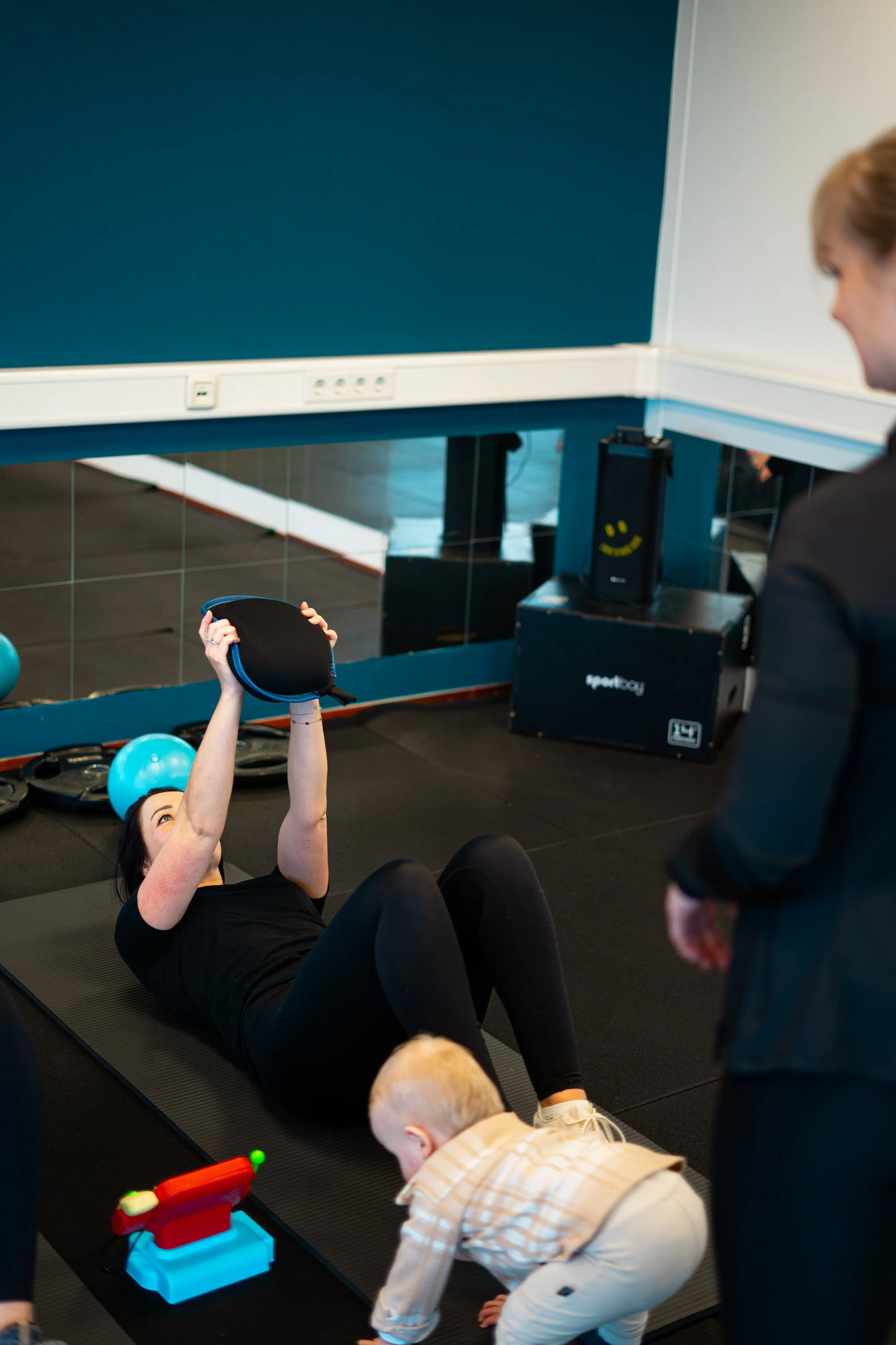 Vrouw ligt op de vloer in de sportschool met een gewicht in haar handen, terwijl een kind ernaast op de grond speelt.