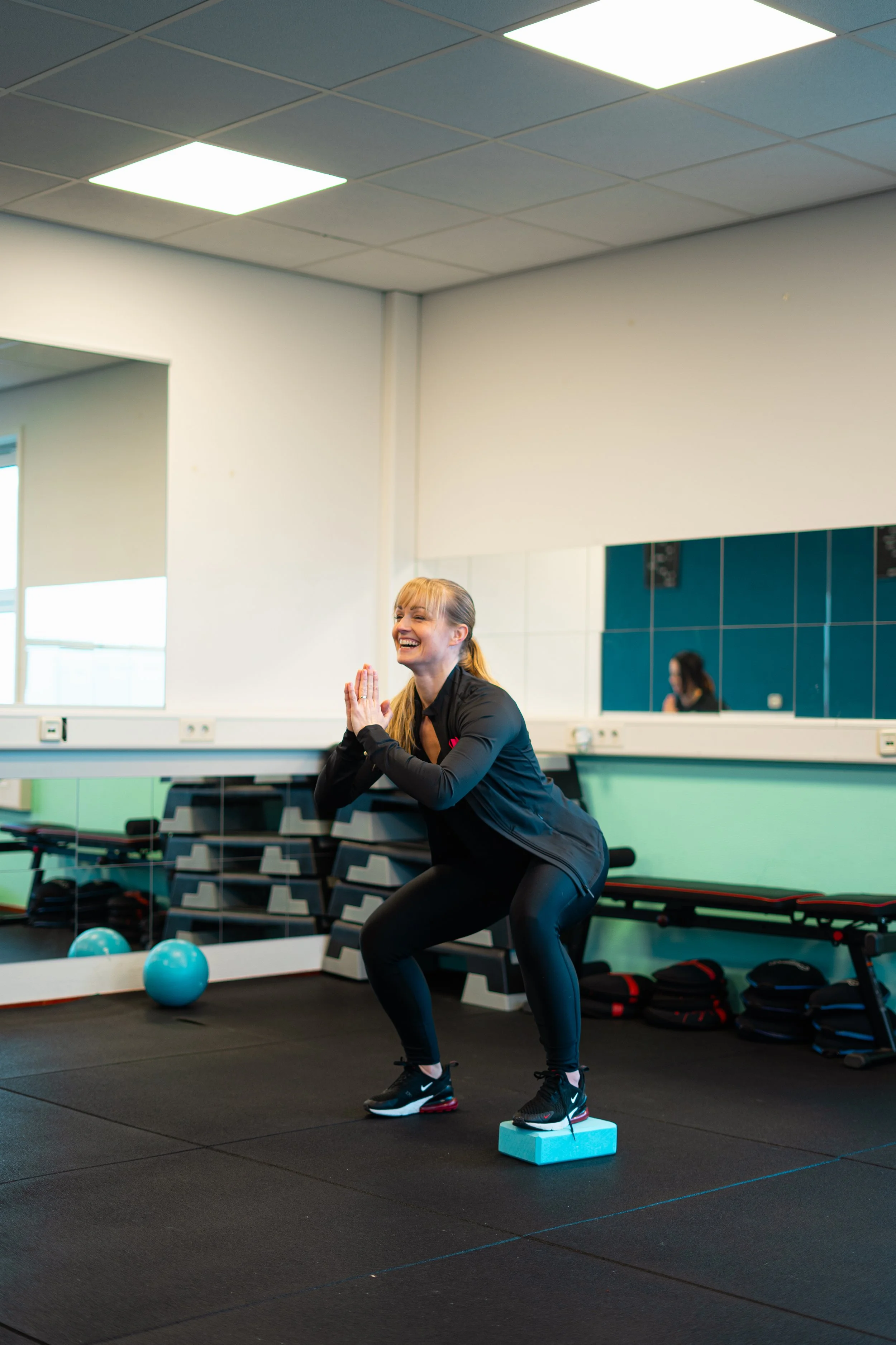 Vrouw die een squat oefening doet in een sportschool, met een foamblok onder haar voeten, lachend en in sportkleding.