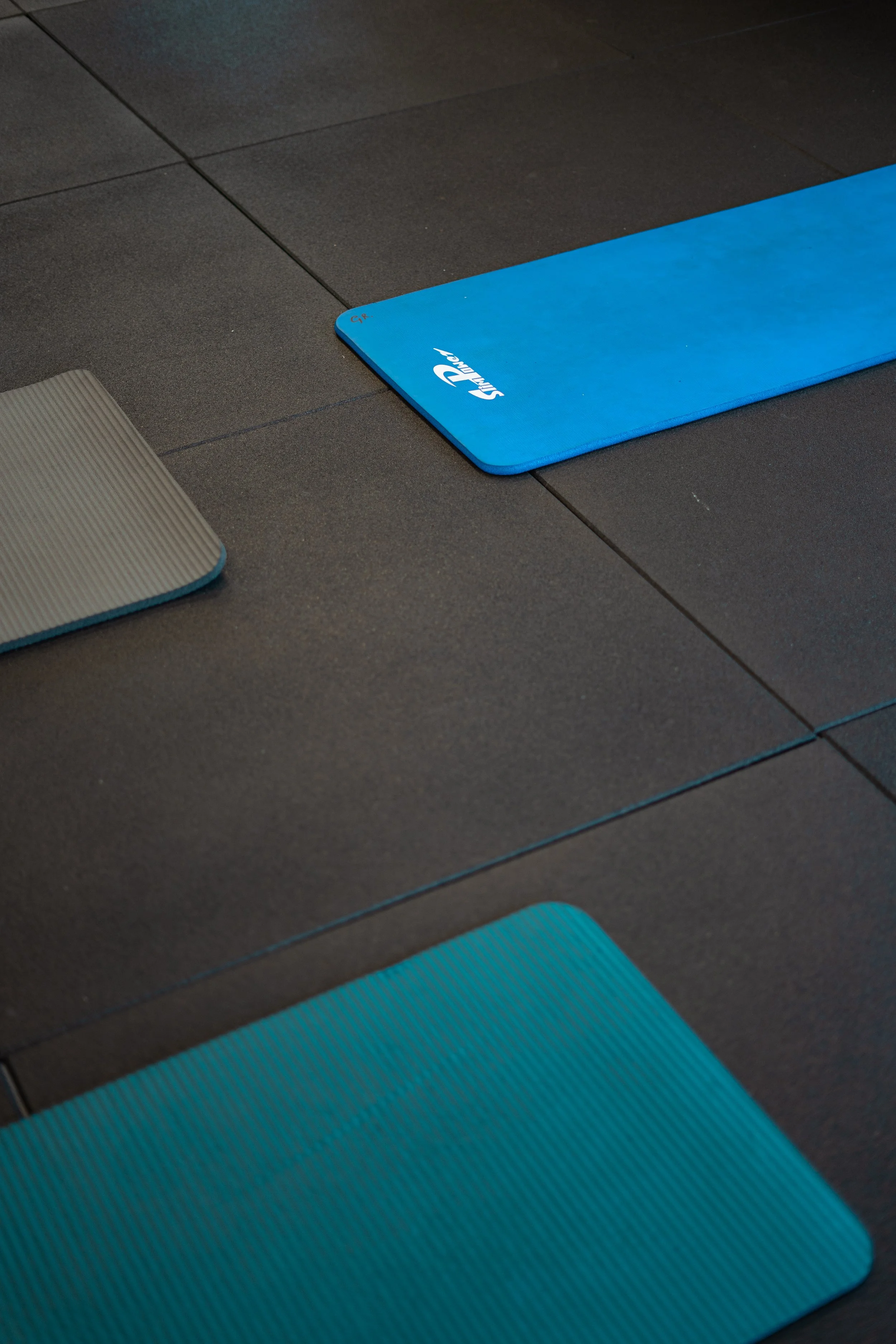 Drie fitnessmatten liggen op een zwarte vloertegel in een sportschool. Eén mat is grijs met aquablauwe randen, een andere is helemaal blauw met een logo dat 'SILVER' zegt, en de derde is blauw met een ribbelstructuur.
