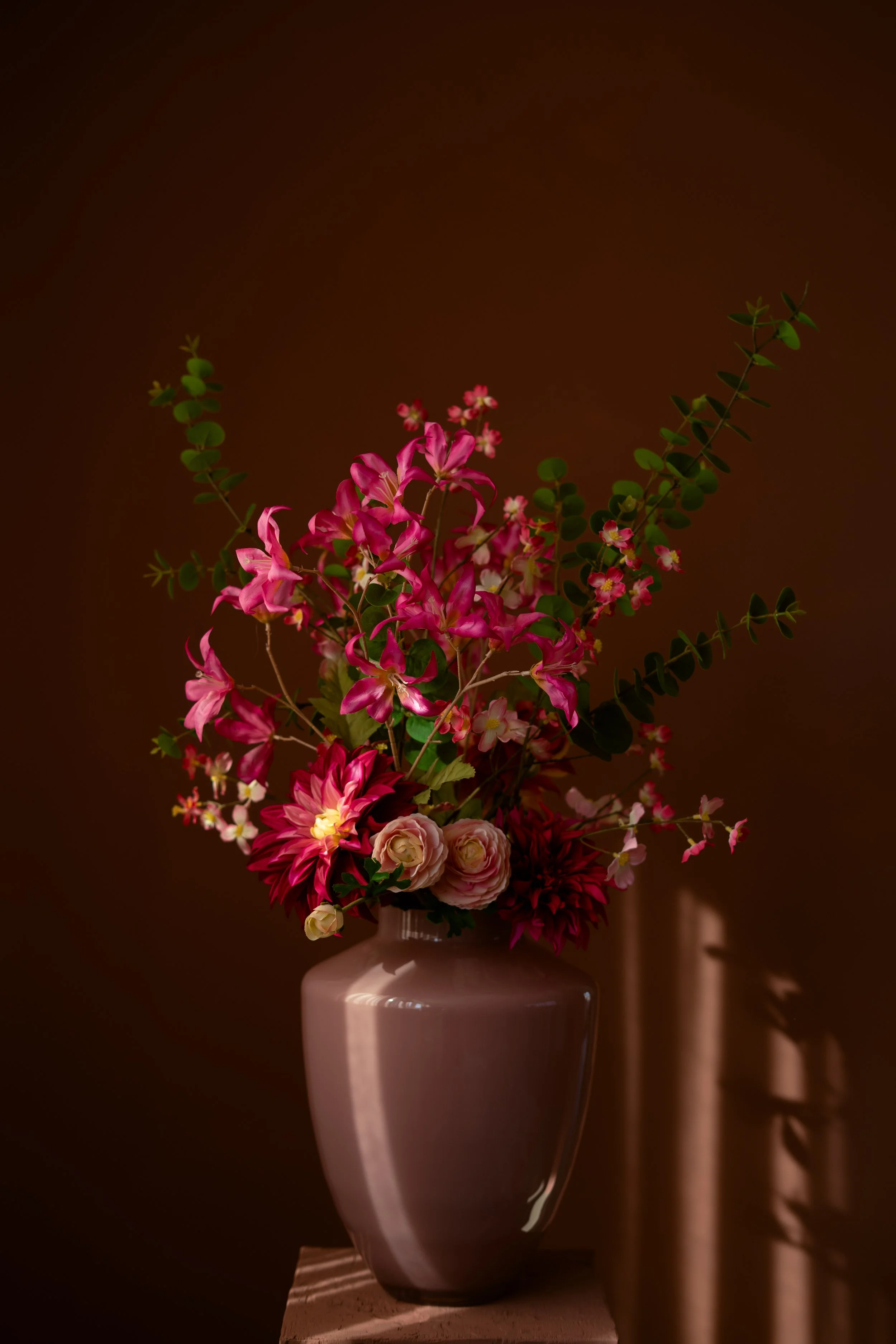 Een bloemstuk met verschillende roze en rode bloemen in een matte roze vaas, geplaatst op een sokkel met een donkere achtergrond. Floweris