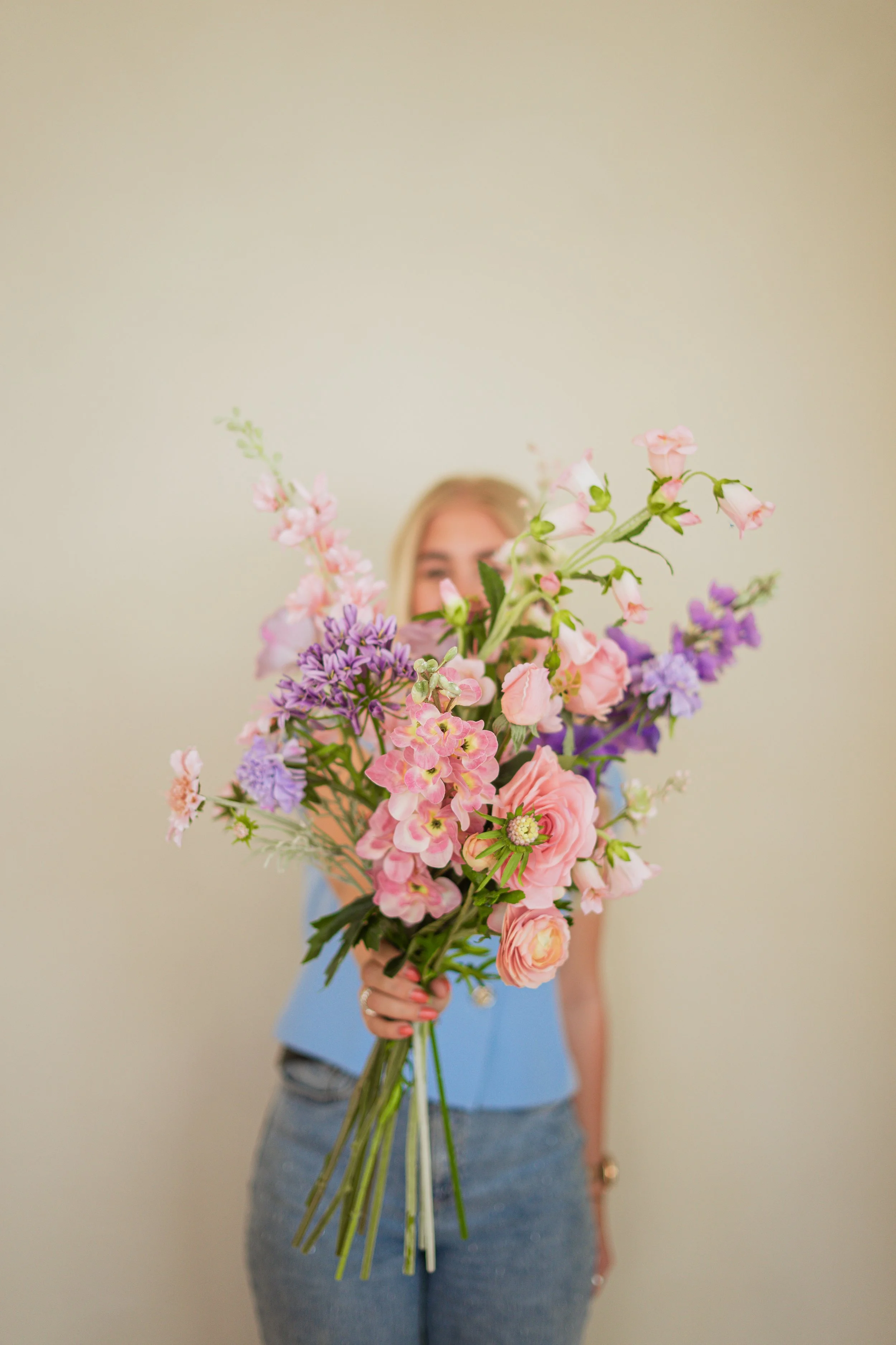 Vrouw houdt een grote bos bloemen voor zich, bloeiend met roze en paarse bloemen in een kamer met een lichtgele muur. Floweris