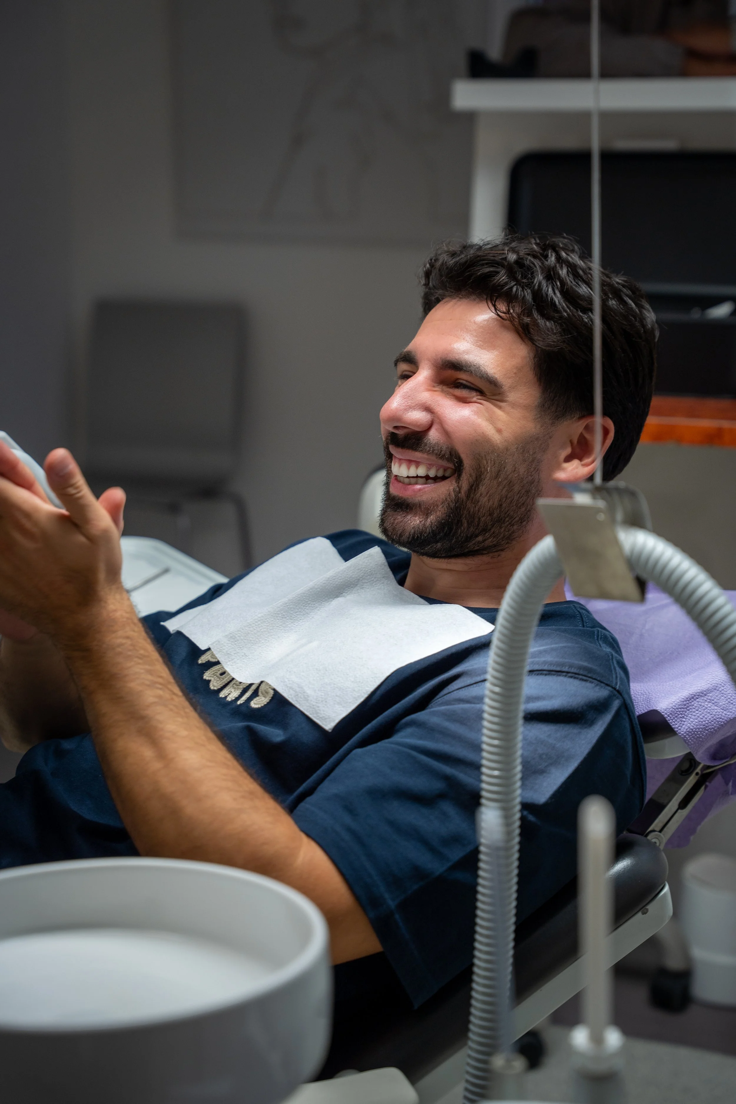 Man ligt op een tandartsstoel, lachend, met een beschermende cape en een papieren doekje. Aan de rechterkant is een tandartsapparaat zichtbaar in een tandartspraktijk. Wittewel. Tandenbleken.