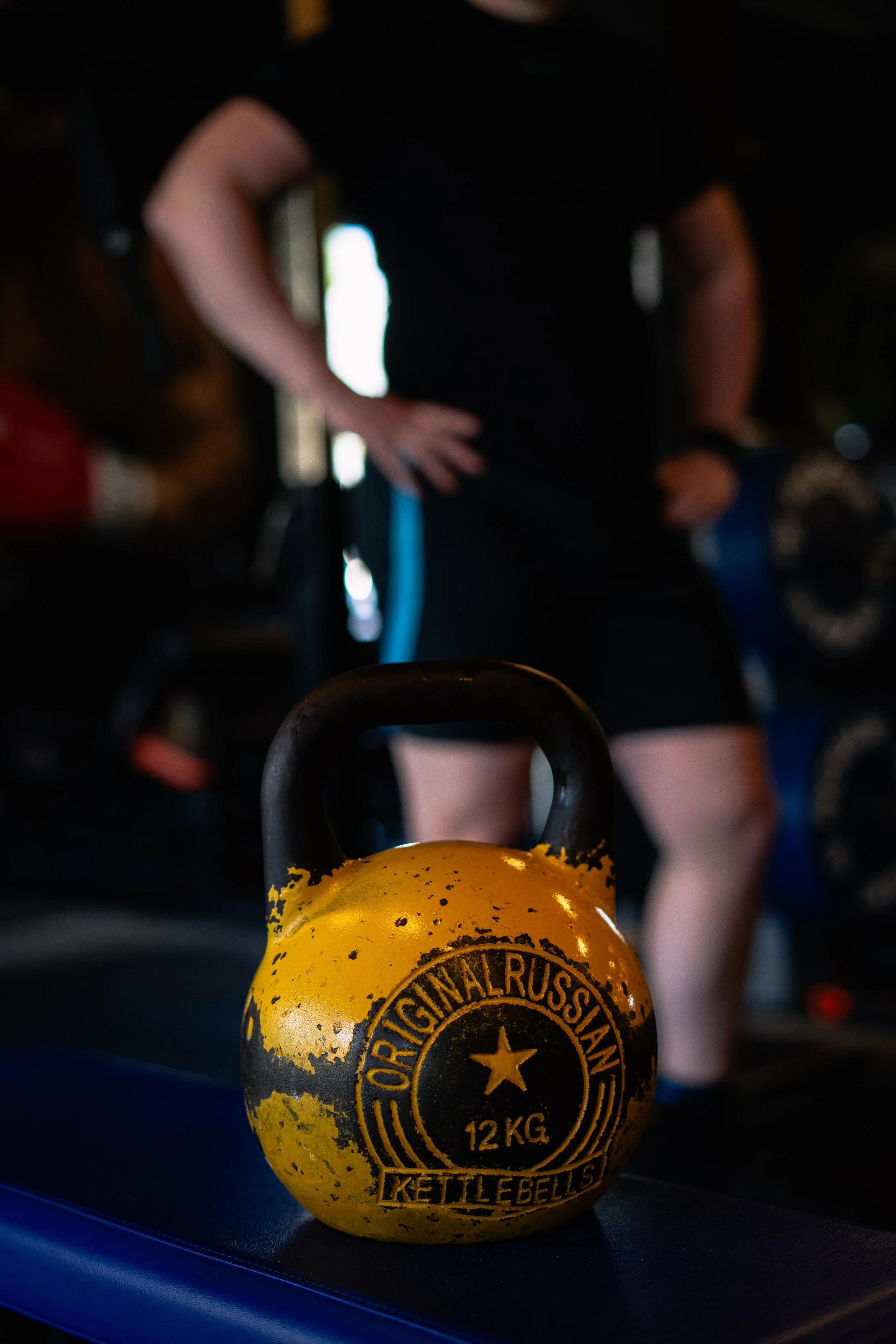 Een gele kettlebell met zwarte tekst ligt op een loopband, terwijl een persoon in de achtergrond aan het trainen is in een fitnessruimte. Sportschool Emmen. Krachttraining.