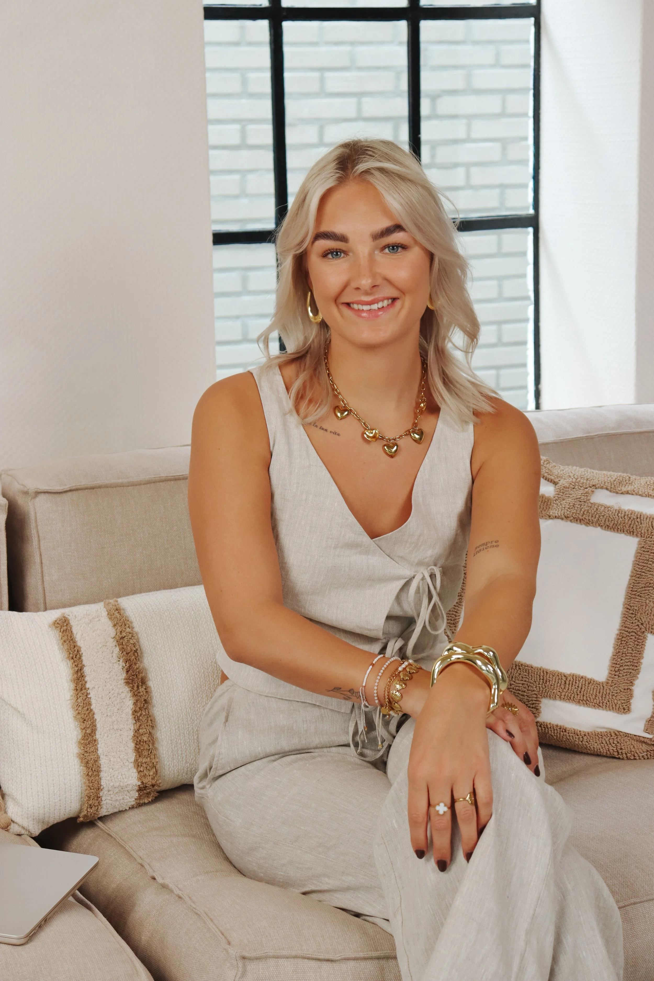 Een jonge vrouw met blond haar, blauwe ogen en een glimlach, zittend op een bank in een interieur met grote ramen en een witte muur. Branding shoot