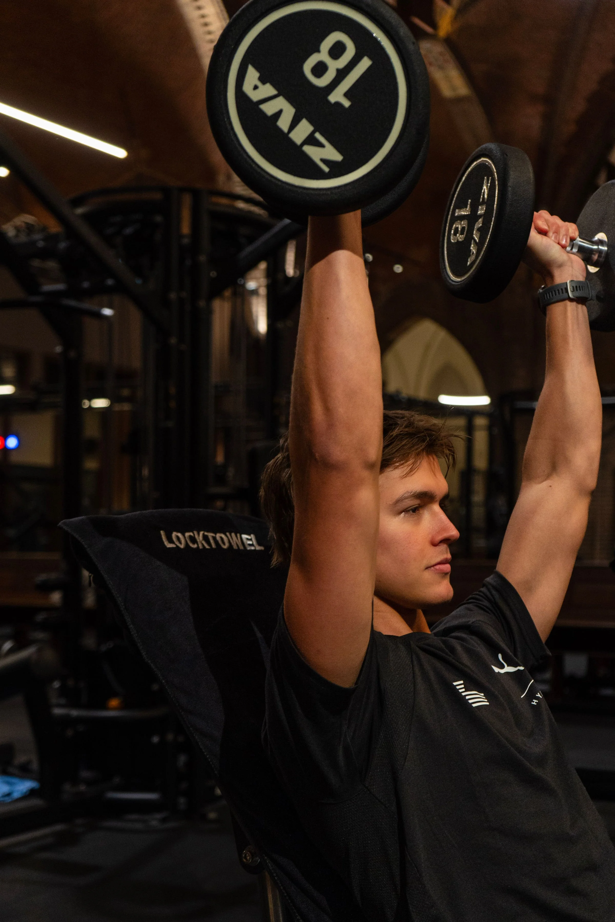 Een jonge man traint in een sportschool en tilt twee dumbbells boven zijn hoofd, met de achtergrond van fitnessapparatuur en een houten plafond. Sporthanddoek. Locktowel.