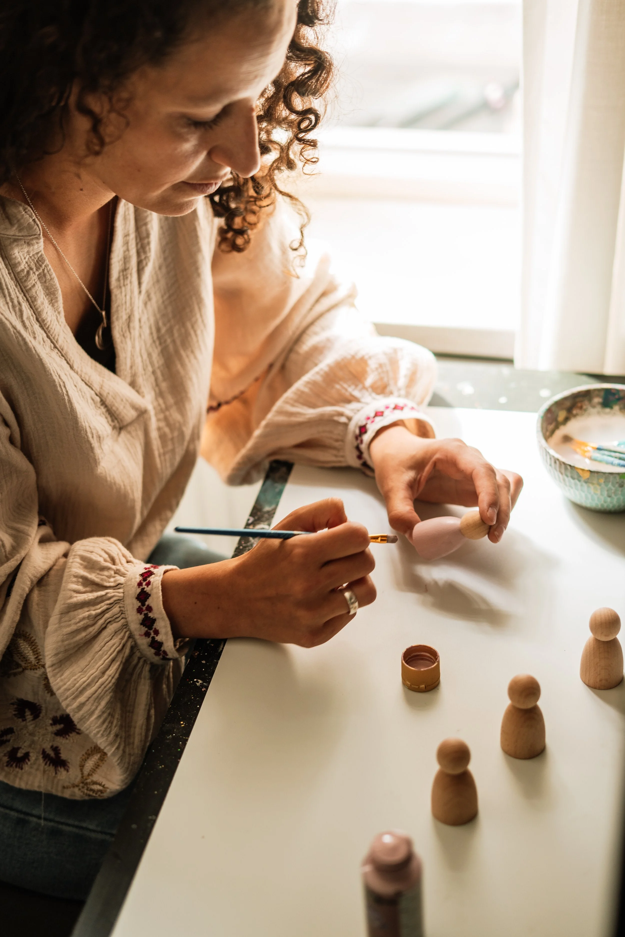Vrouw schildert een houten pop op een witte tafel in een lichte kamer bij een raam.
