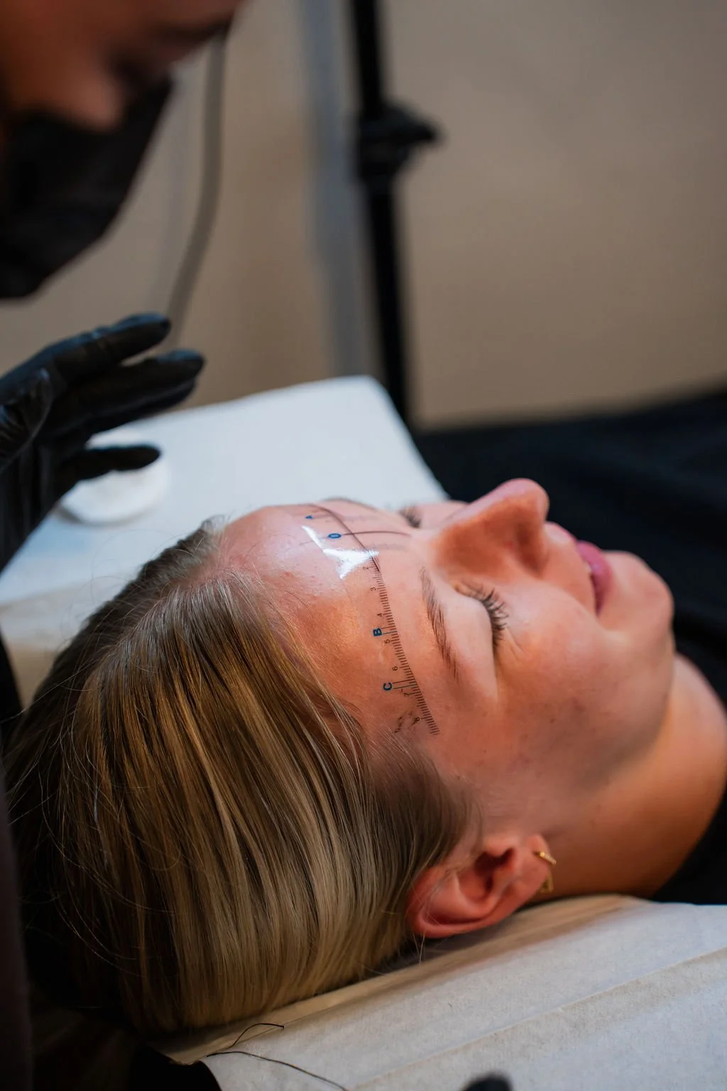 Een vrouw ligt op haar rug met een meetlint boven haar voorhoofd, terwijl een arts of specialist een procedure uitvoert. De vrouw heeft blonde haren en draagt een klein oorbel. De specialist draagt zwarte handschoenen en een masker. Beautysalon.