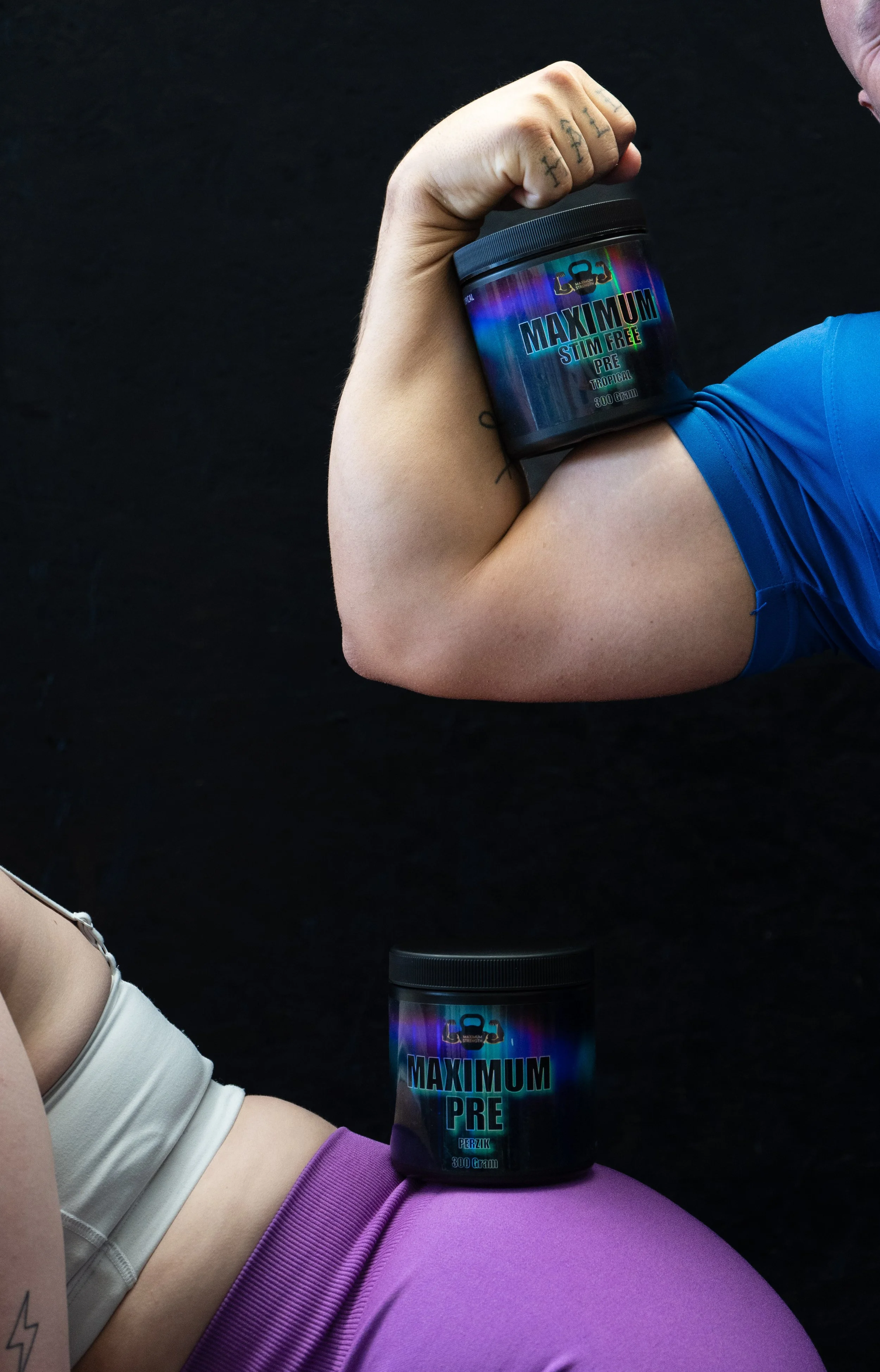 Een persoon die zijn gespierde arm toont met een container van Maximum Stim Free pre-workout supplement dicht bij zijn biceps. Andere persoon heeft een container op hun dij en draagt roze sportlegging en een beige sporttop.