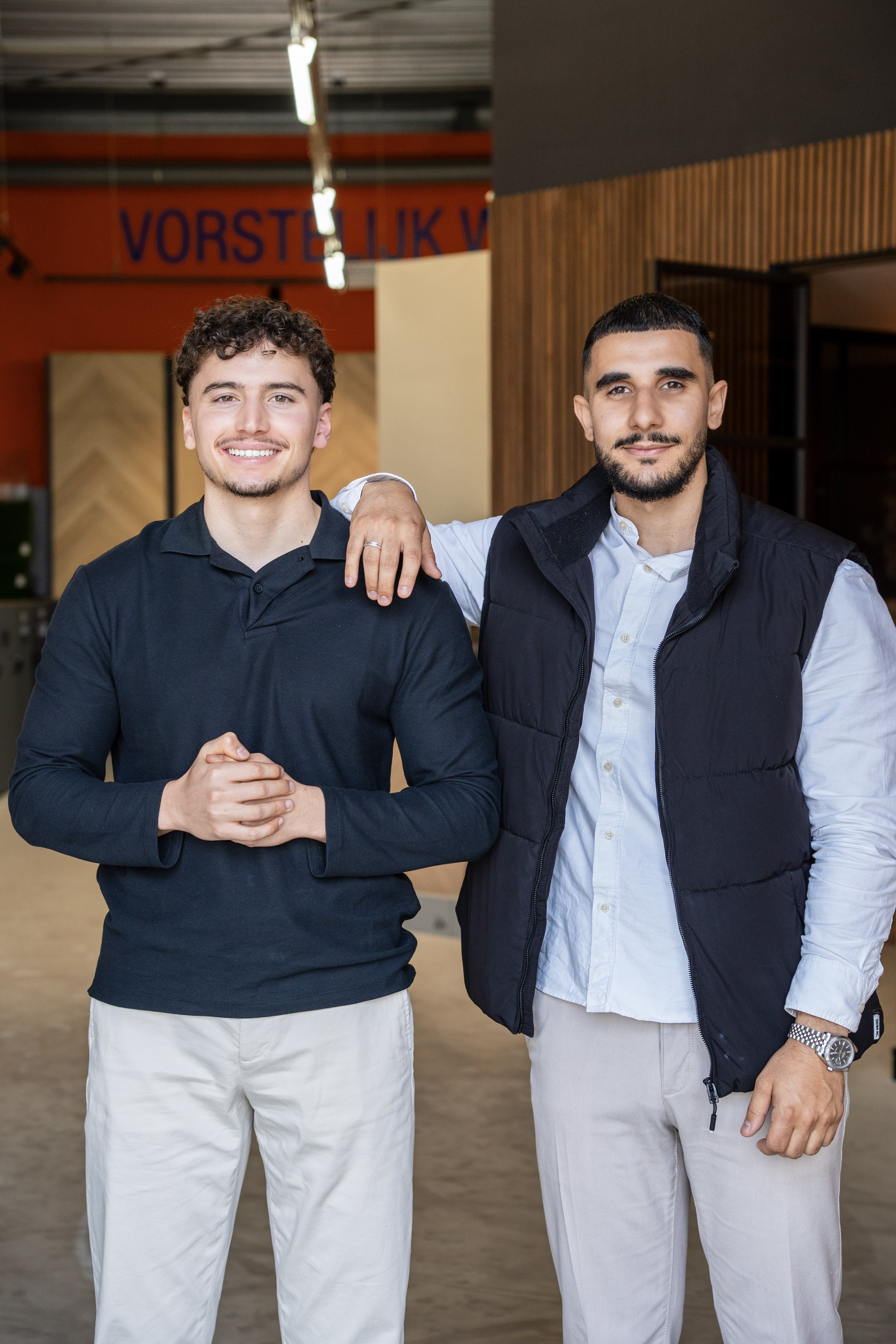 Twee jonge mannen die samen poseren in een binnenruimte, de ene met een arm om de schouder van de andere, beiden glimlachend. Laminaatpaleis Ede