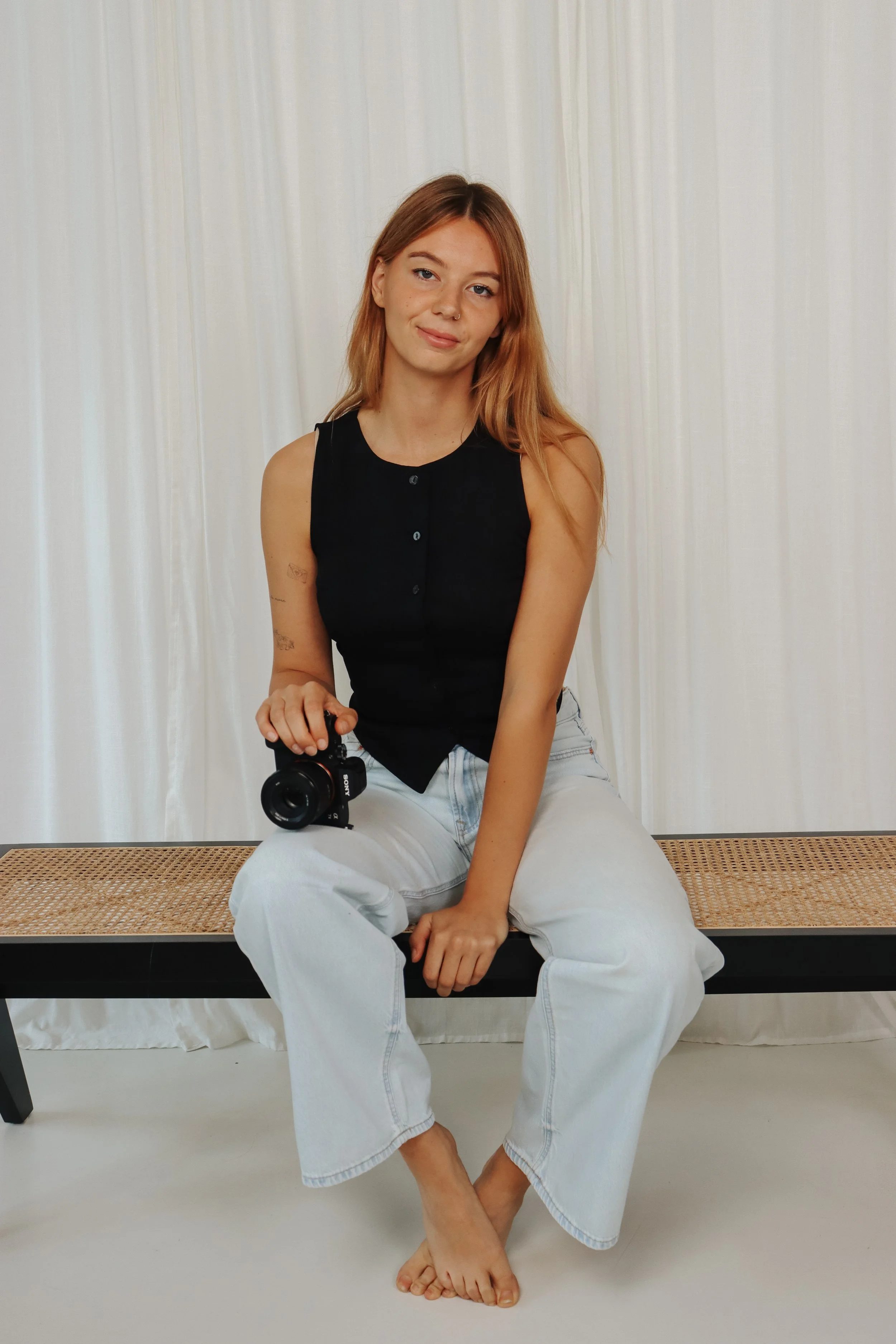 Een jonge vrouw met rood haar zit op een bank, draagt een zwarte top en witte wijde jeans, houdt een camera in haar rechterhand, en zit met haar benen gekruist op de vloer voor een lichte gordijnachtergrond. Branding shoot