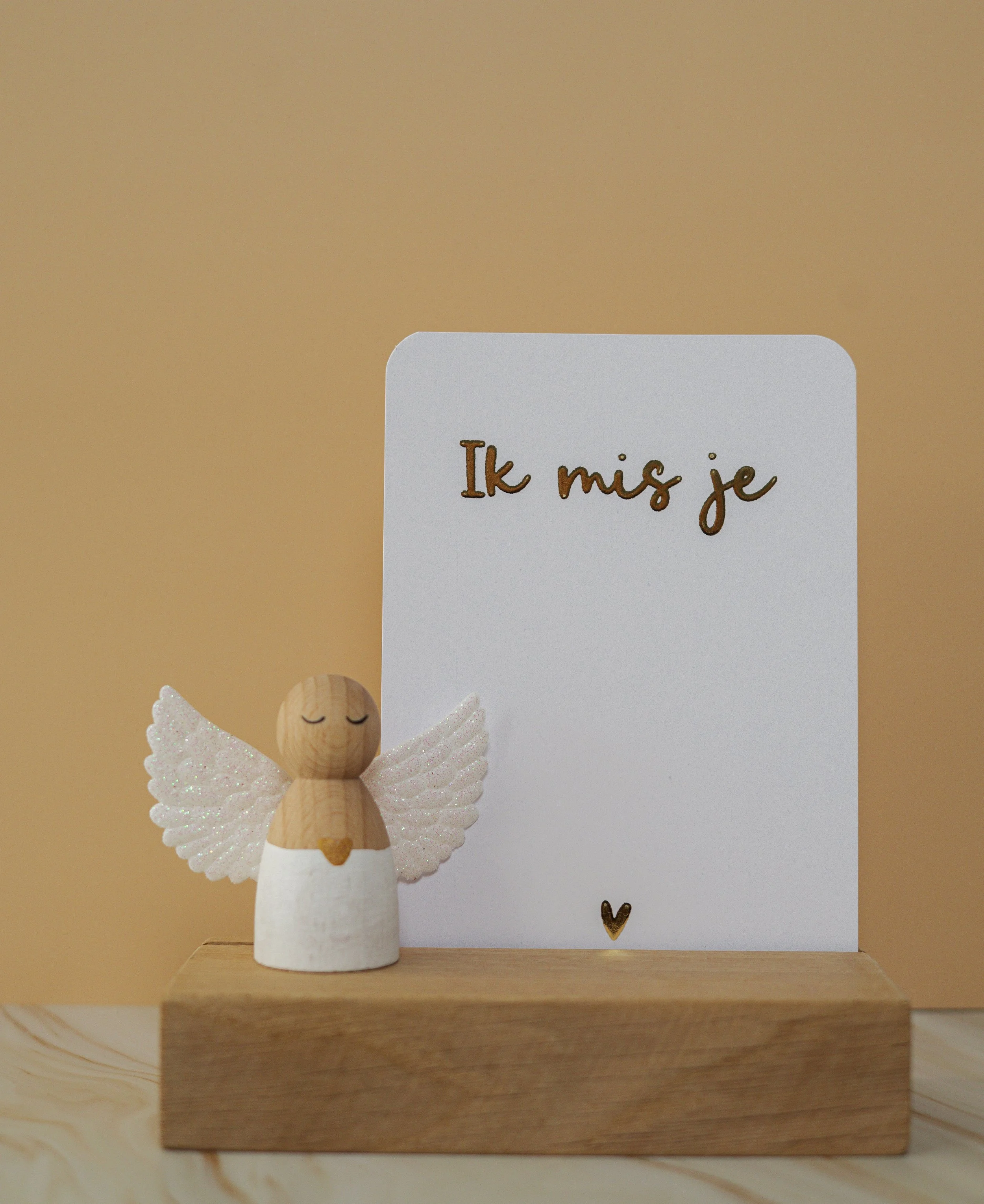 Een klein houten engeltje met witte sierlijke vleugels staat naast een wit kaartje waarop staat 'Ik mis je' met een klein goudkleurig hartje eronder, op een houten standaard.