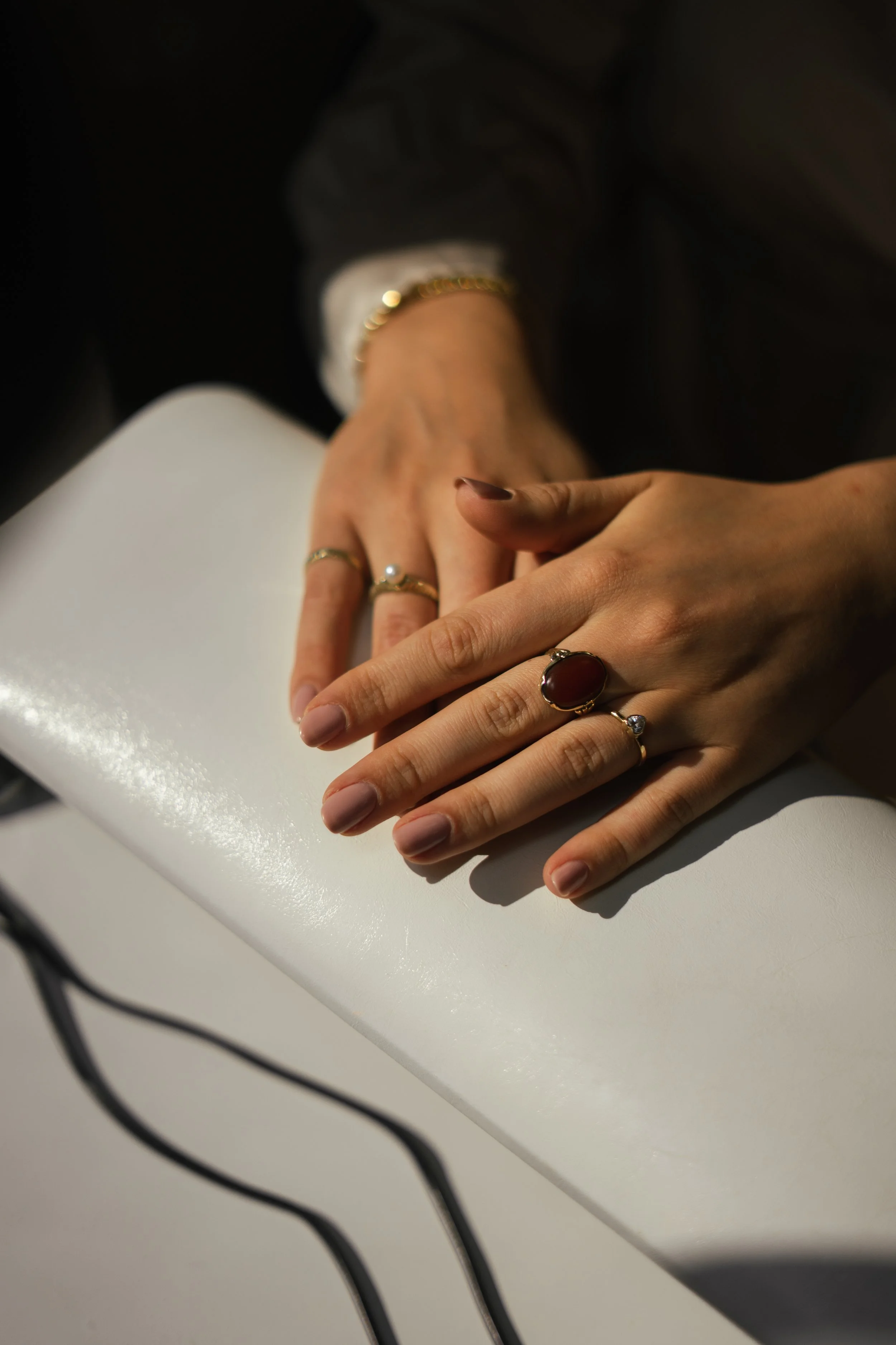 Een close-up van twee handen die op een witte tafel rusten, met verschillende sieraden zoals ringen en een armband, onder helder licht.