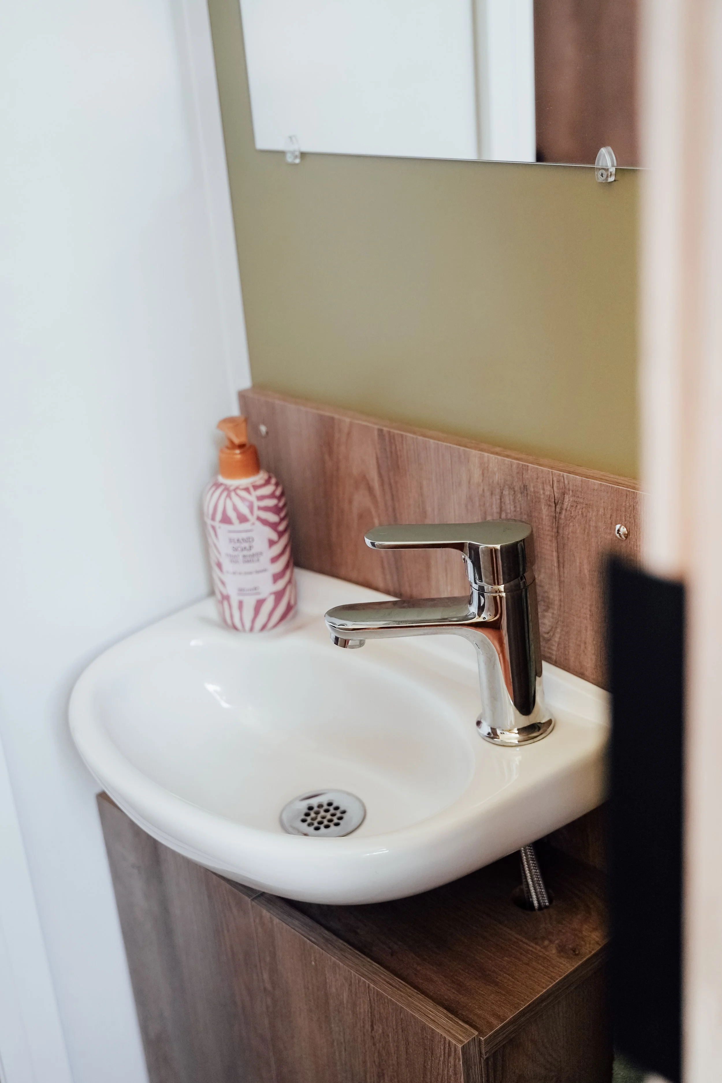 Een klein wit wastafel met een moderne chroomkraan, een fles handzeep met roze en witte patroon op de rand, een houten muurafwerking en een spiegel. Vakantiepark. Chalet.