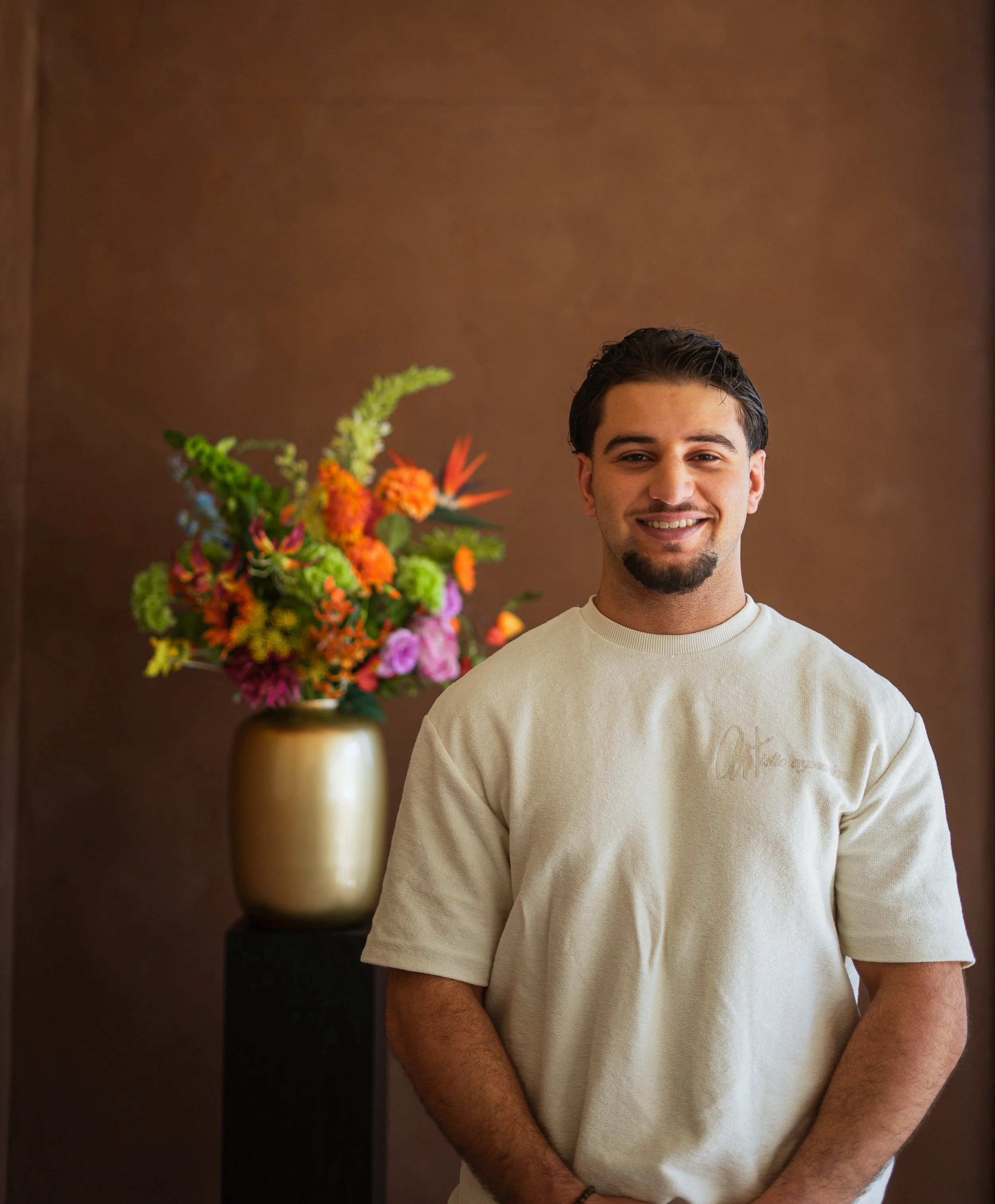 Een lachende man met een beige T-shirt staat voor een bruine achtergrond en een grote bloemstuk in een gouden vaas. Floweris