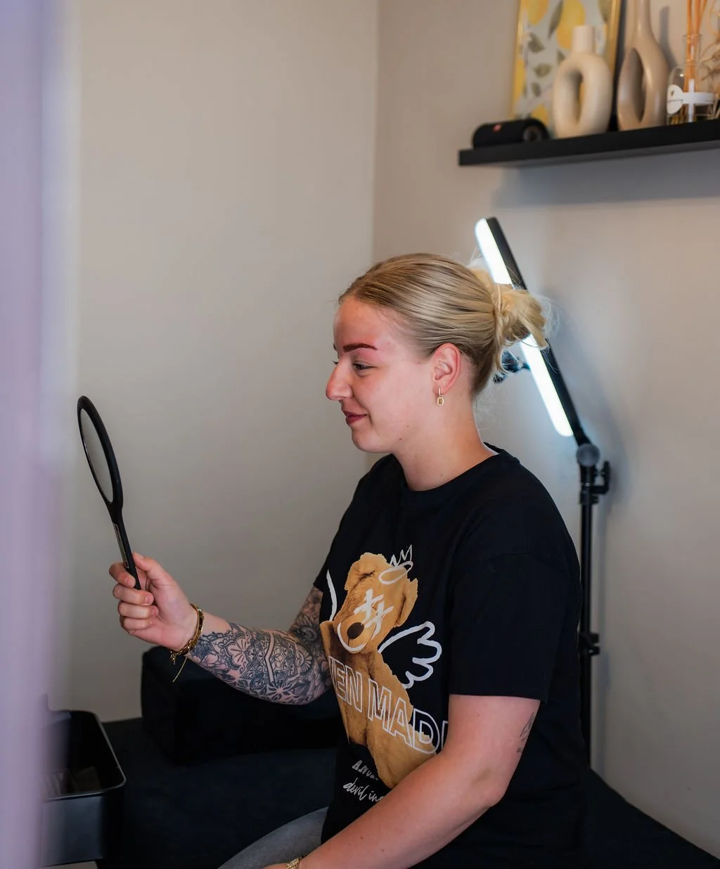 Vrouw met geverfd wenkbrauwen en tatoeages op arm, die naar haar spiegelbeeld kijkt met een glimlach, in een kamer met een zwarte bedrand en een fotolijst en decoratieve vazen op een plank. Beautysalon.