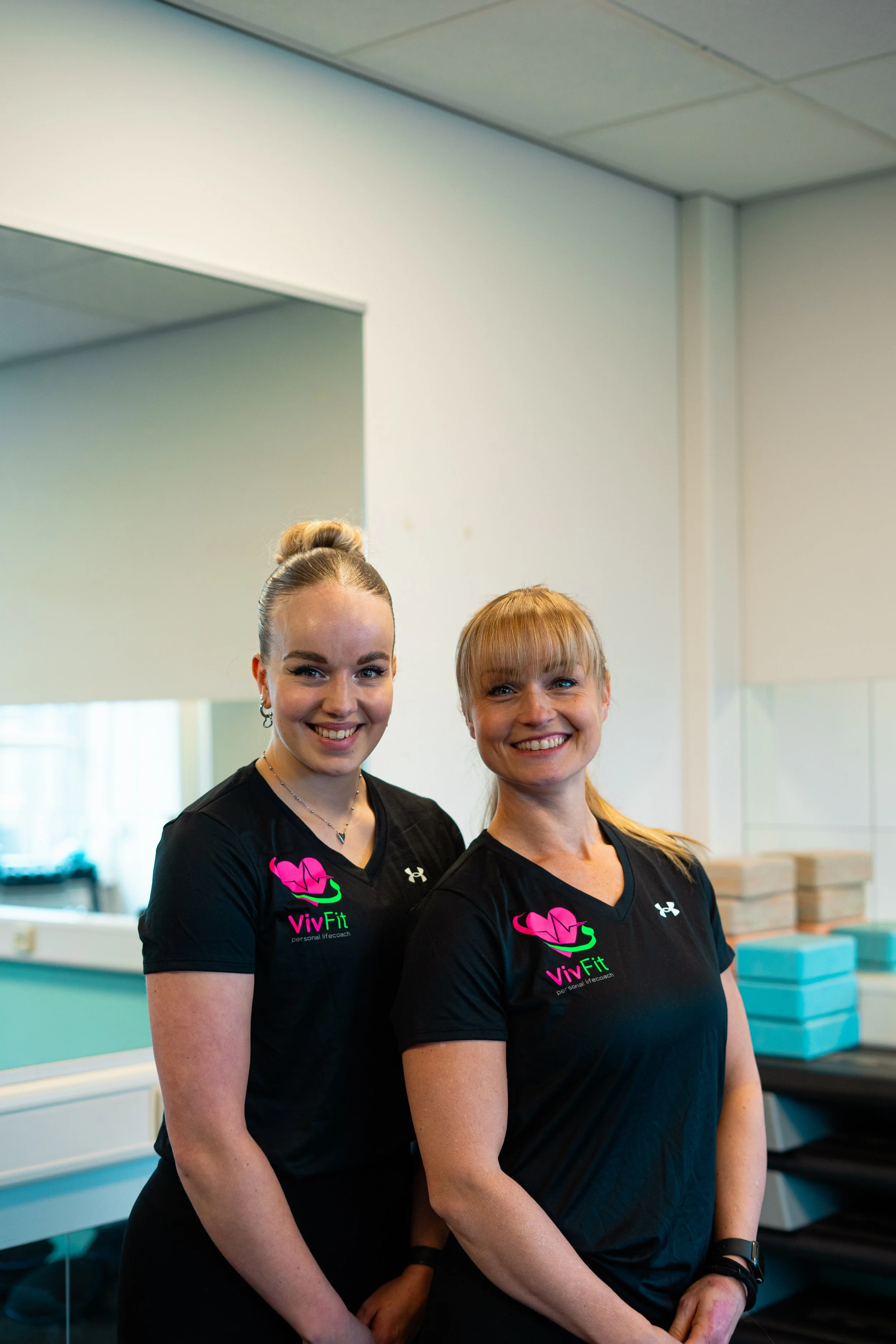 Twee vrouwen in fitnesskleding met het VivFit-logo, glimlachend in een fitnessruimte.