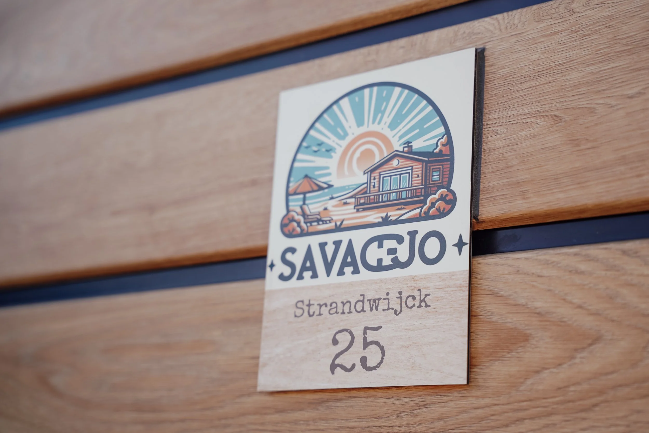Een borden met een ontwerp van een strandhuis, zon en zee, en tekst 'SAVAGEJO' en 'Strandw Jck 25'. Vakantiepark. Chalet.