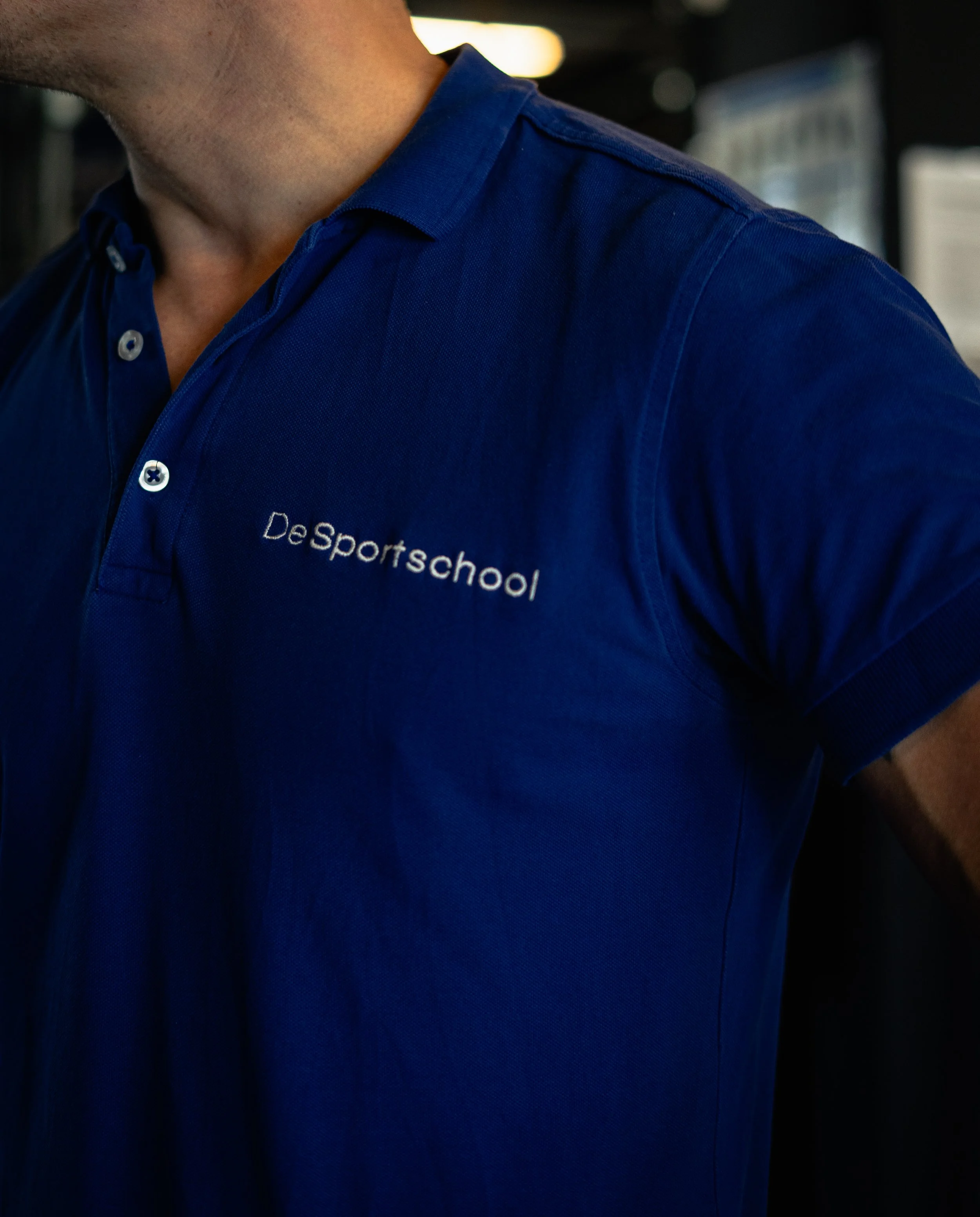 Close-up van iemand in een blauw polo met witte knopen en de tekst 'DeSportschool' op de linker borst.